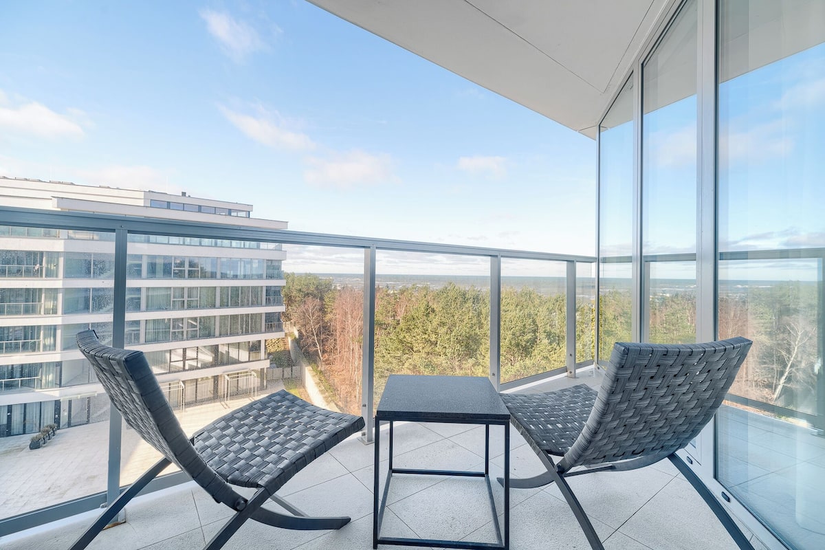 Ein Balkon mit zwei Korbstühlen und einem kleinen Tisch, mit Blick auf eine Stadtlandschaft und Grünflächen.