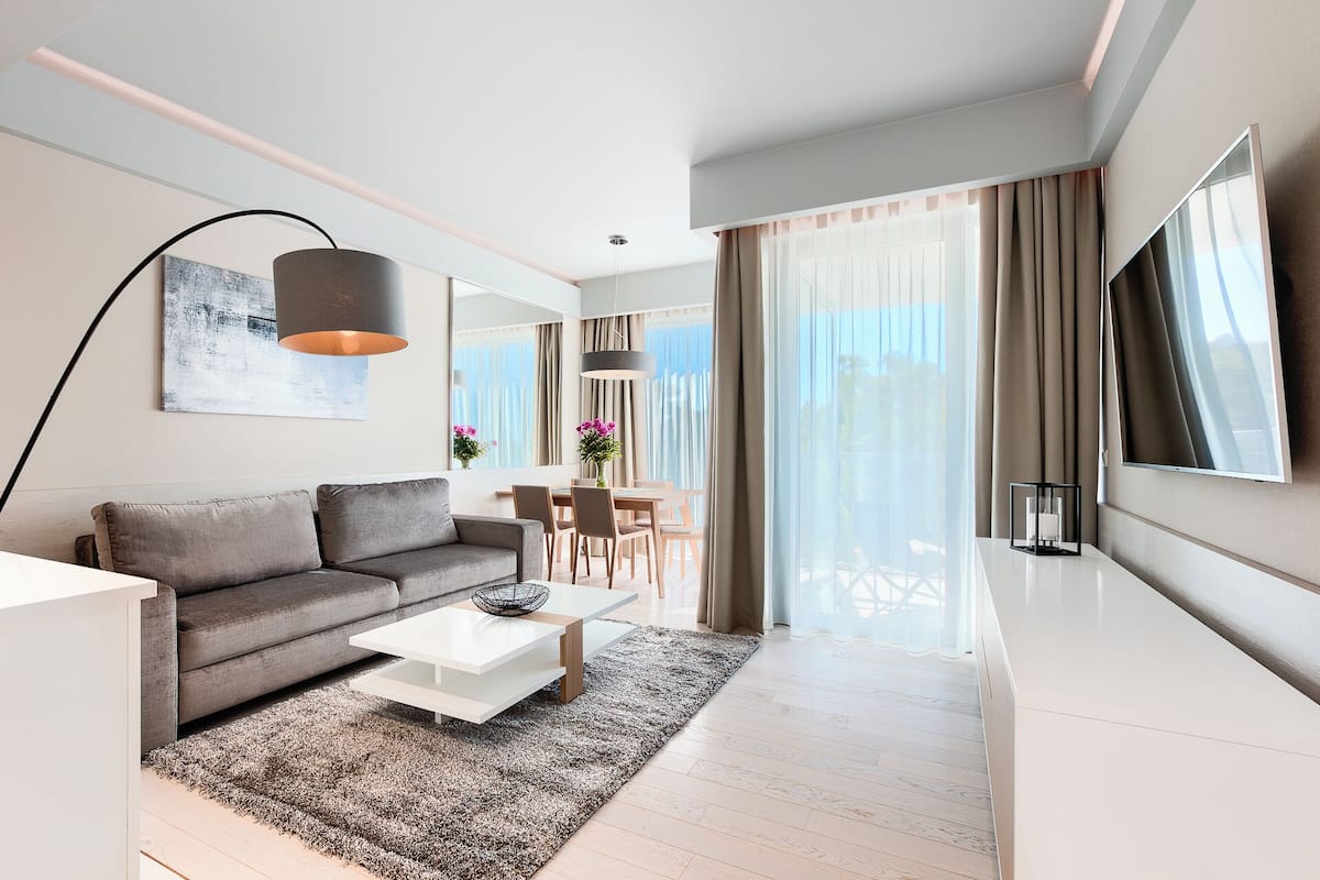 Ein modernes Wohnzimmer mit grauem Sofa, weißem Couchtisch und einem großen Fernseher. Es gibt einen Essbereich mit Tisch und Stühlen sowie einen Balkon mit Vorhängen.