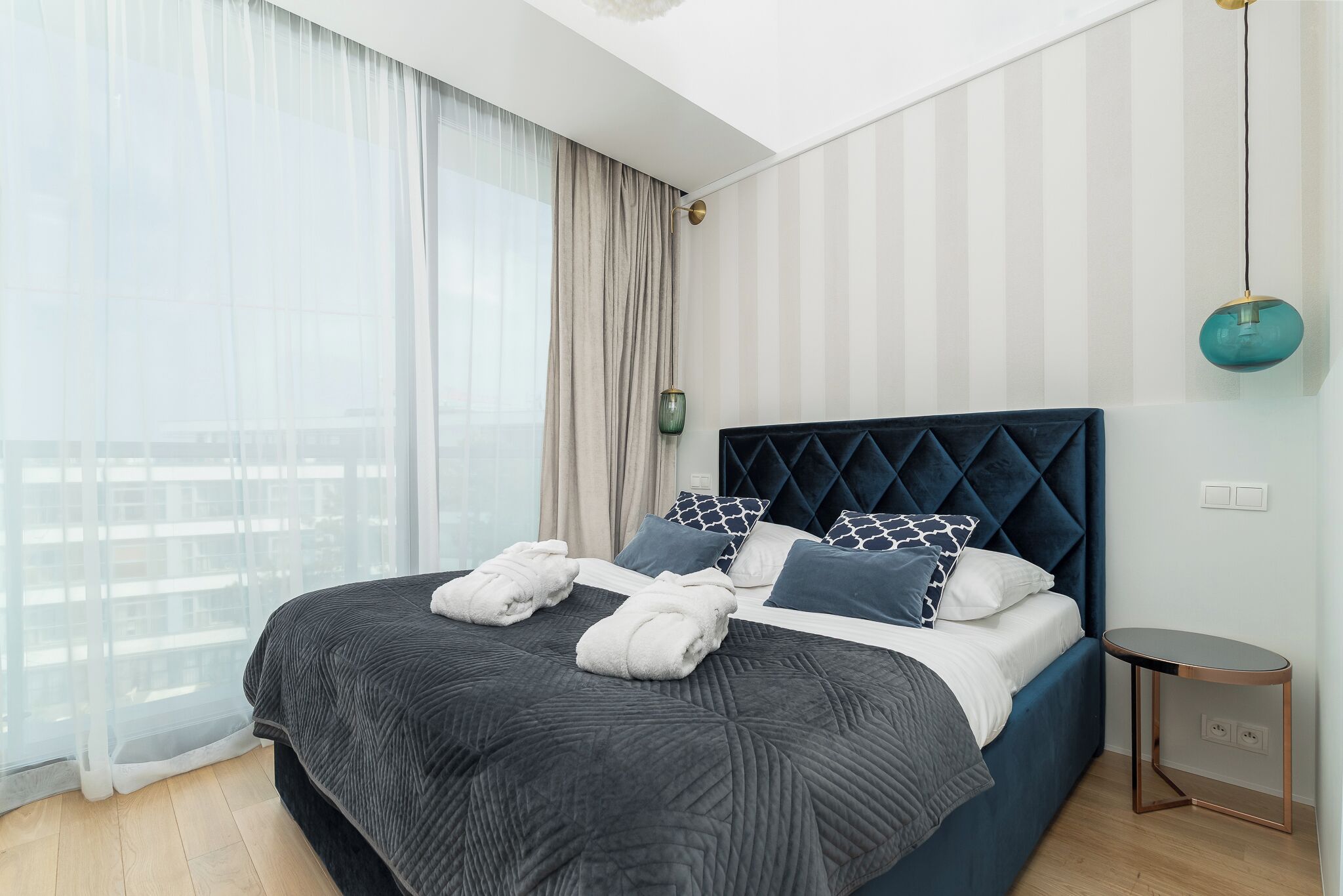 Une chambre d’hôtel moderne avec un grand lit, une table de chevet, un luminaire suspendu et une vue à travers des rideaux transparents.