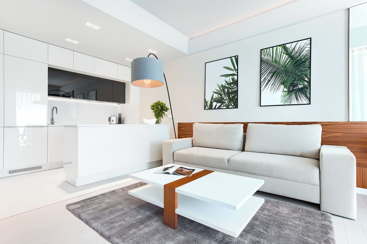 Ein modernes Wohnzimmer mit einem weißen Sofa, einem Couchtisch und einer Küchenzeile mit weißen Schränken und einem grauen Teppich.