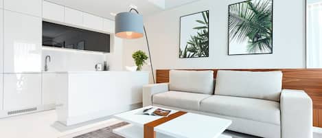 A modern living room with a white sofa, a coffee table, and a kitchen area with white cabinets and a grey rug.