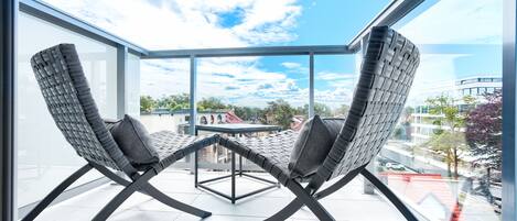 Un balcon avec deux chaises et une table, de grandes fenêtres et une vue sur les immeubles et les arbres.