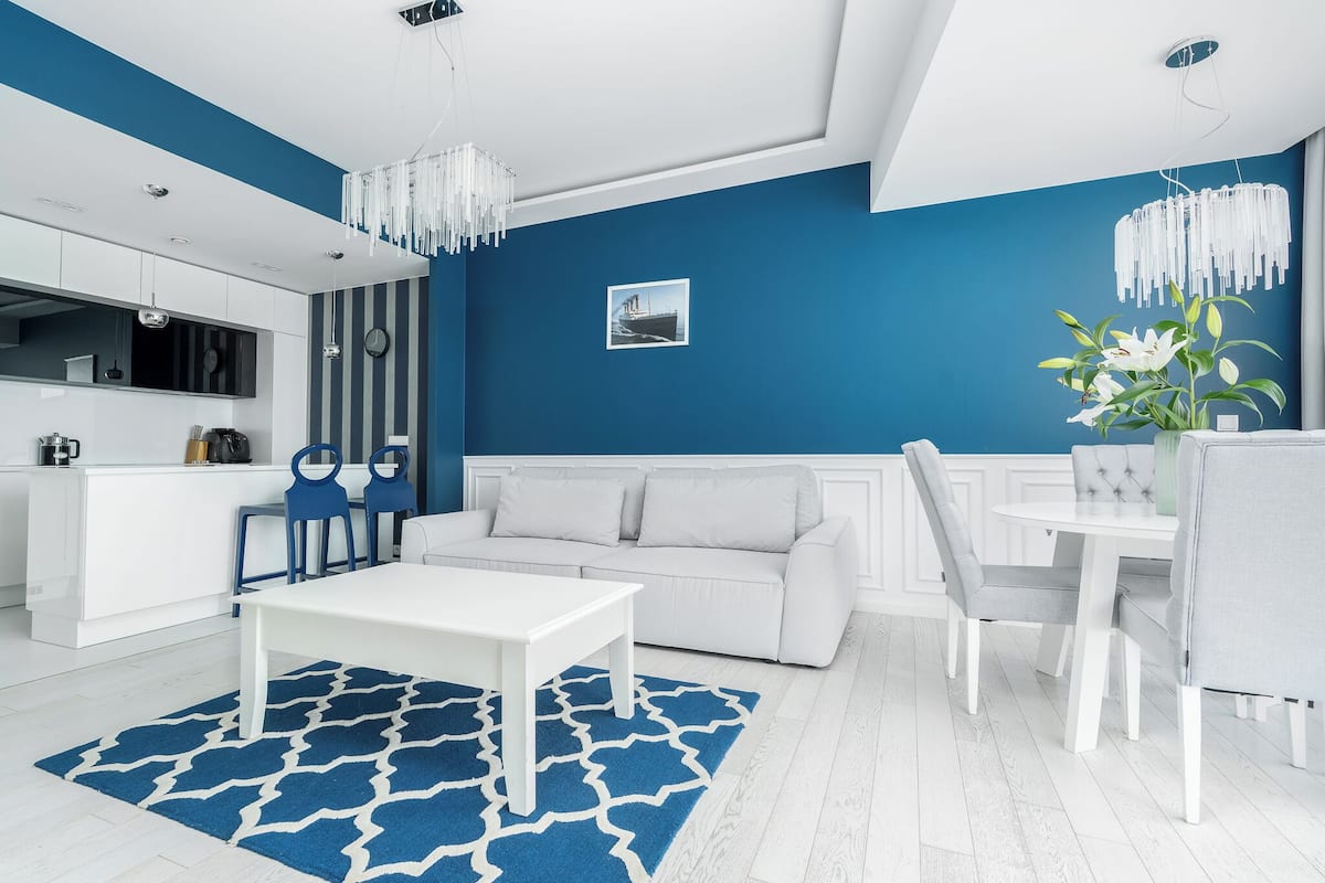 Ein modernes Wohnzimmer mit einem weißen Sofa, einem Esstisch und Stühlen. Es gibt eine Küchenzeile mit weißen Schränken und einer blauen Akzentwand.