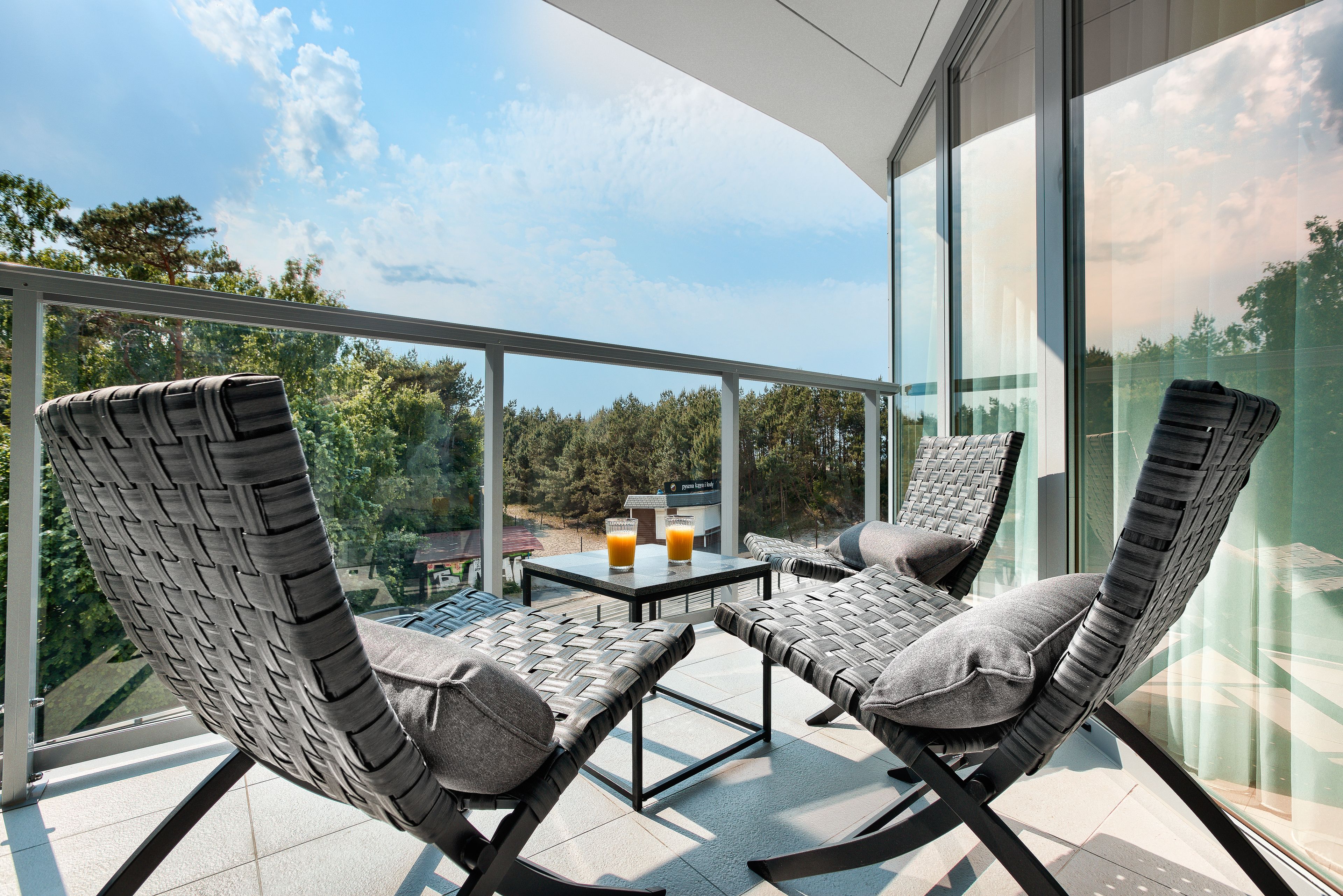 A balcony with wicker chairs and a table, two glasses of orange juice, and a view of trees and buildings.