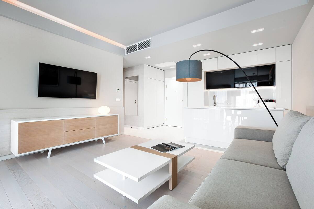 Ein modernes Wohnzimmer mit einem weißen Sofa, einem Couchtisch, einem an der Wand montierten Fernseher und einem durch eine offene Tür einsehbaren Küchenbereich.