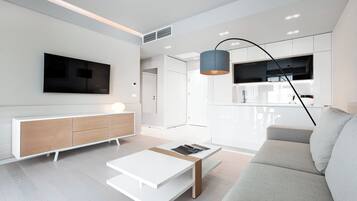 Una sala moderna con un sofá blanco, una mesa de centro, un televisor montado en la pared y una cocina visible a través de una puerta abierta.