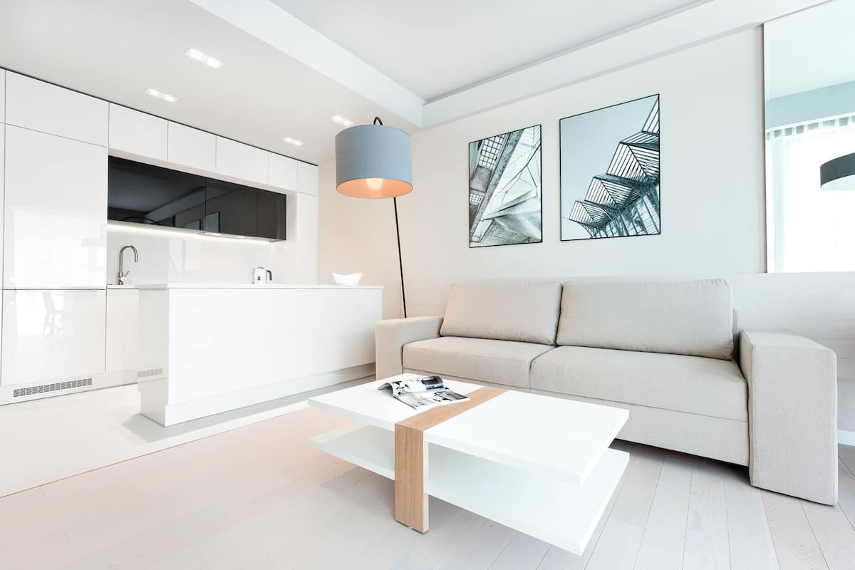 Ein modernes Wohnzimmer mit einem weißen Sofa, einem Couchtisch und einer Küchenzeile mit weißen Schränken und einer Pendelleuchte.