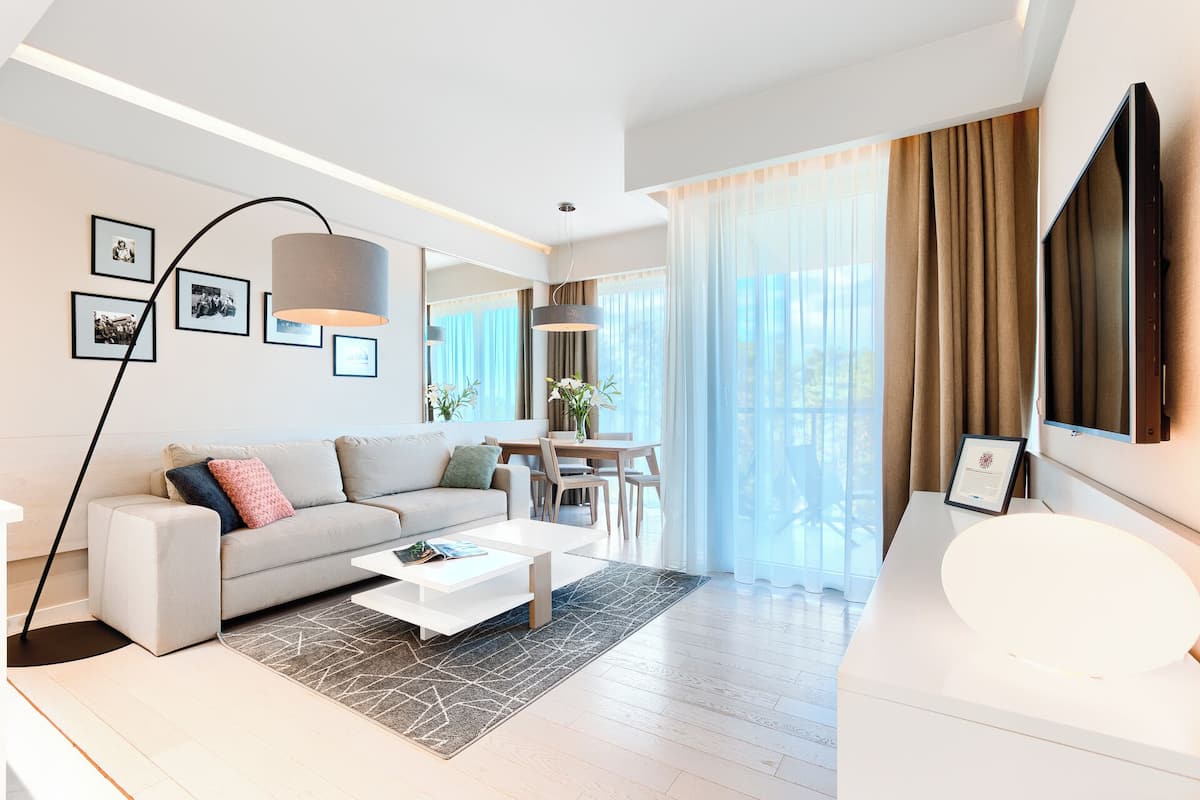 Ein modernes Wohnzimmer mit Sofa, Couchtisch und Essbereich. An der Wand hängen gerahmte Bilder, ein großes Fenster mit Vorhängen und ein flachbildfernseher ist an der Wand montiert.