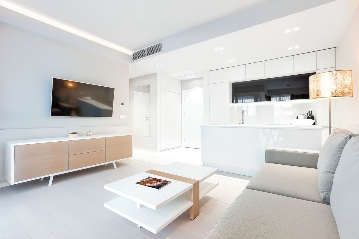 Ein modernes Wohnzimmer mit einem Flachbildfernseher, einem weißen Sofa, einem Couchtisch und einer Küchenzeile mit weißen Schränken und einer Arbeitsplatte.