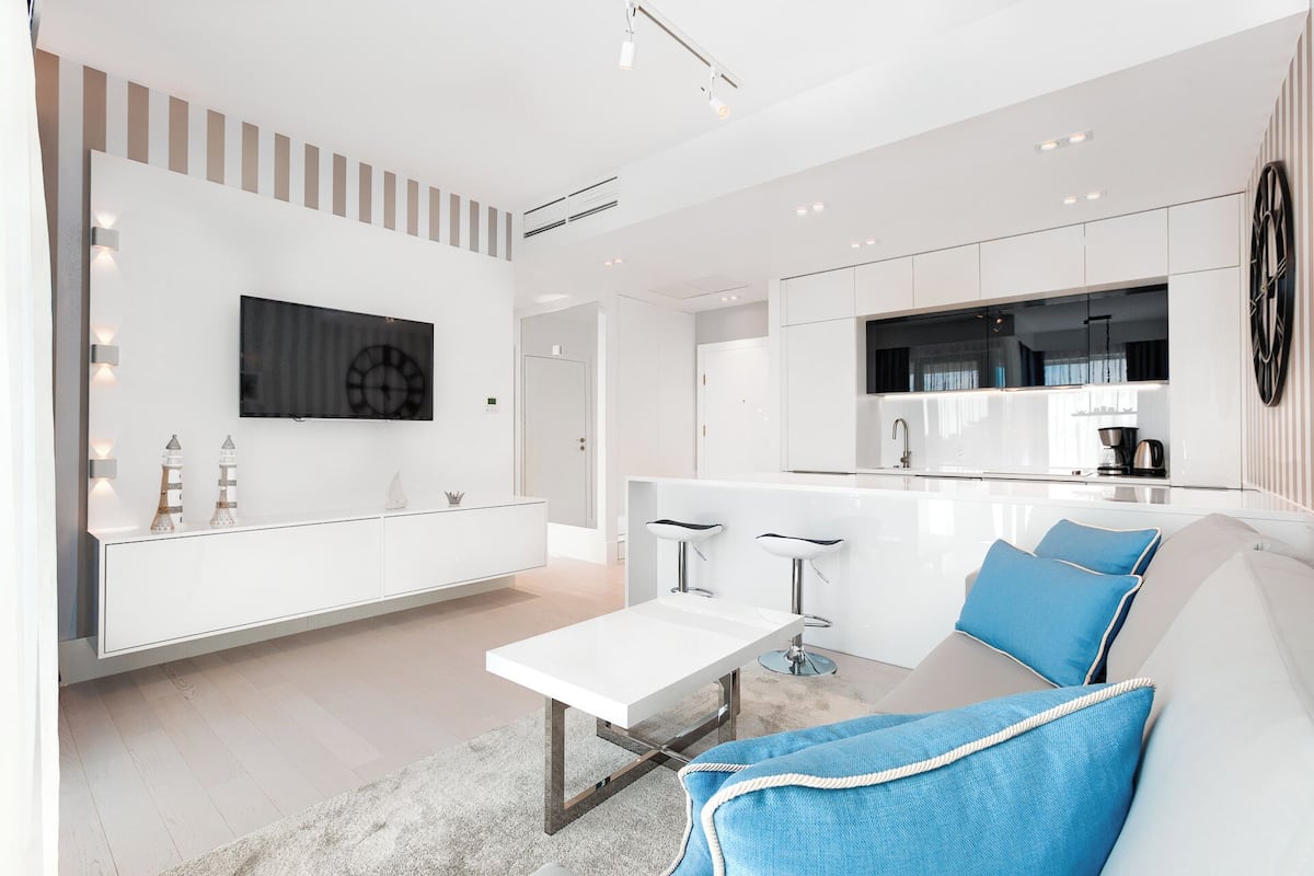 Eine moderne Küche mit einer zentralen Kochinsel, einem Essbereich mit weißem Tisch und einem Wohnbereich mit einer Couch und blauen Kissen.