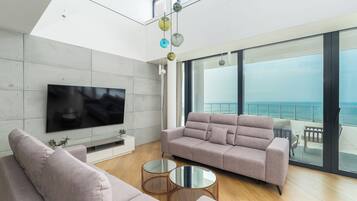 A modern living room with a large flat-screen TV, a sofa, two round coffee tables, and a balcony with a view of the sea.