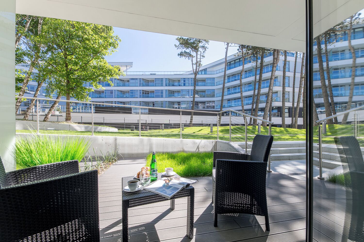An outdoor seating area with wicker furniture, a table with a bottle, and a cup, overlooking a modern building and a grassy area.