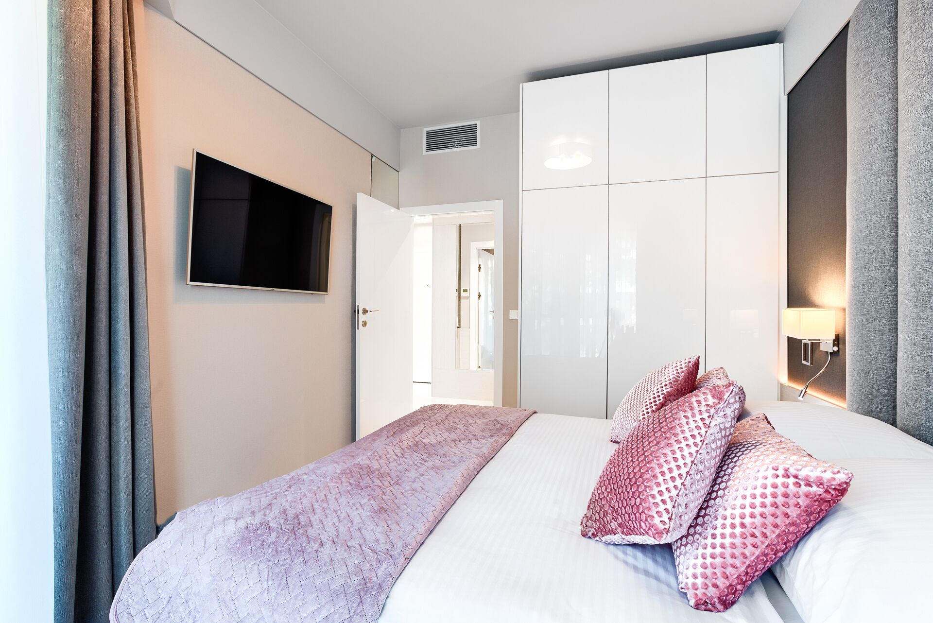 A hotel room with a bed, a TV mounted on the wall, a wardrobe, and a bedside lamp.