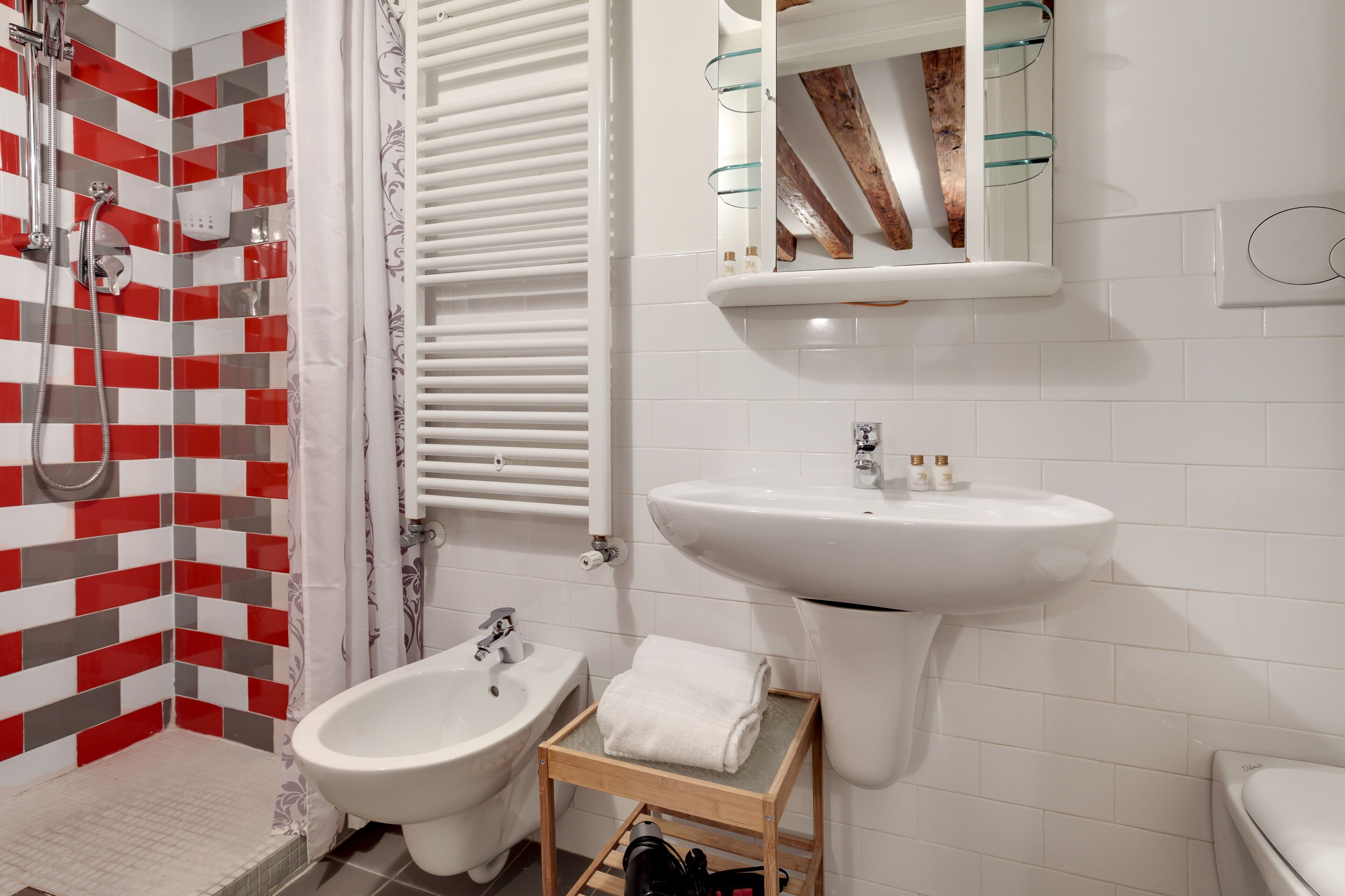 A bathroom with a shower, bidet, sink, and a mirror with a wooden beam.