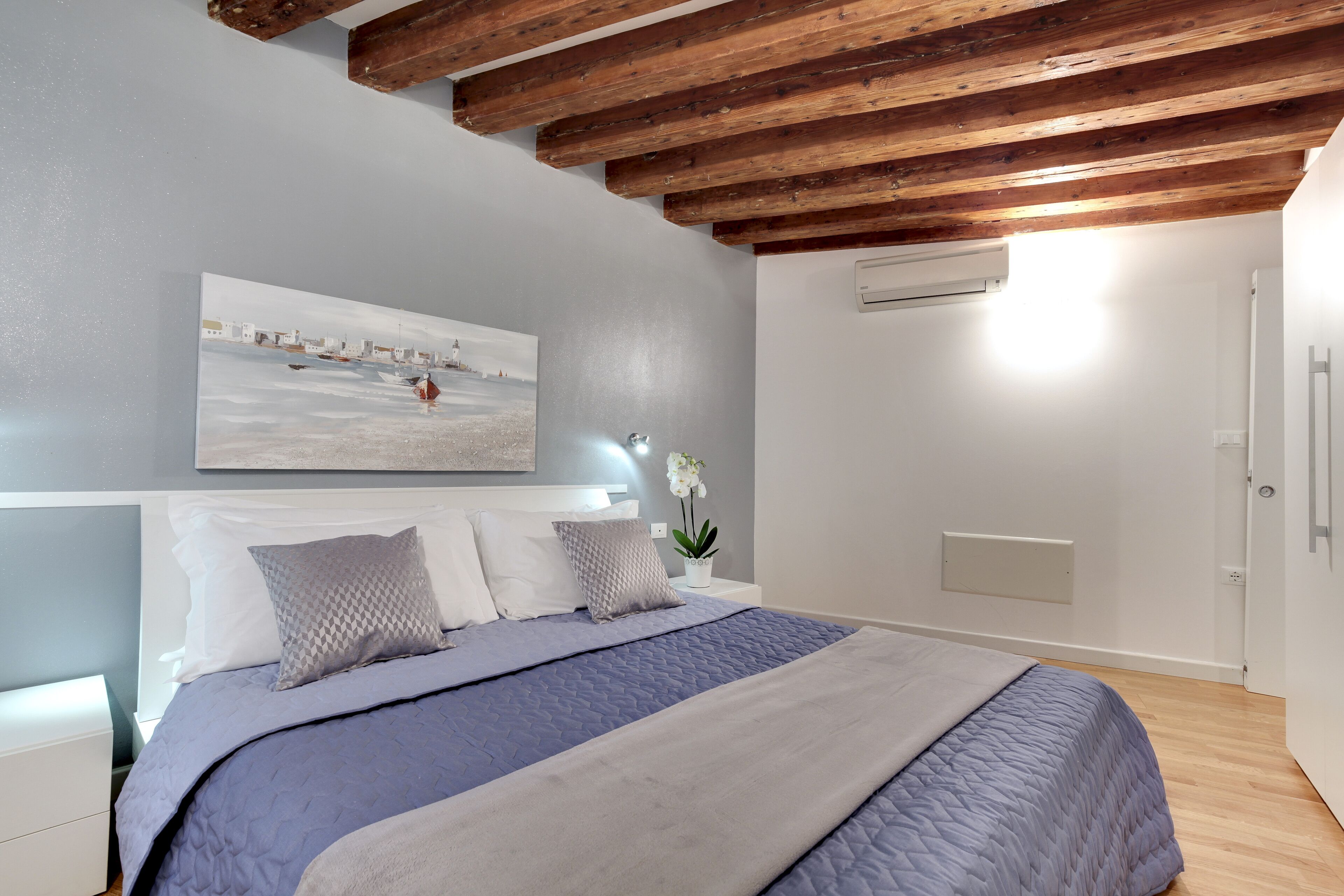 A bedroom with a bed, a nightstand, a wall-mounted air conditioner, and a painting of a coastal scene.