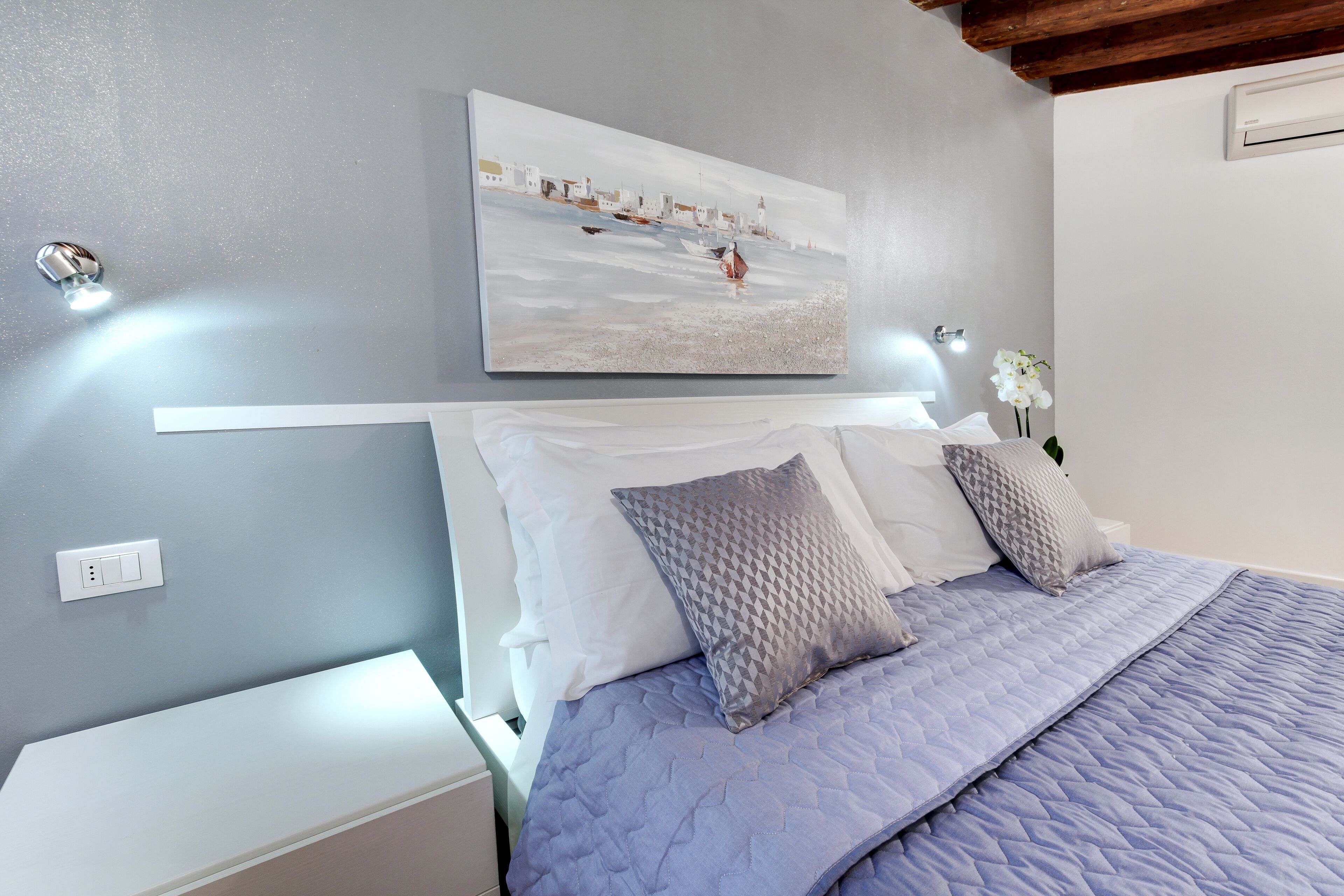 A bedroom with a bed, white nightstands, a wall-mounted painting, and a white flower vase.