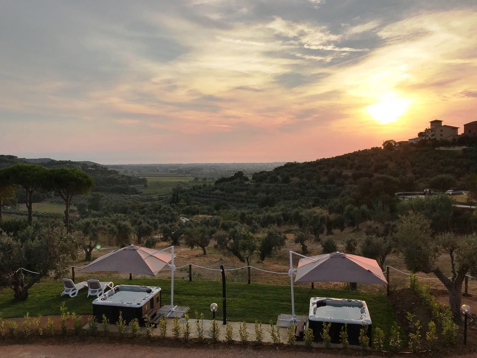 Vista panorámica de un paisaje campestre con un atardecer, que incluye un jacuzzi y sillones de descanso bajo sombrillas, rodeado de olivos y una colina distante con edificaciones.