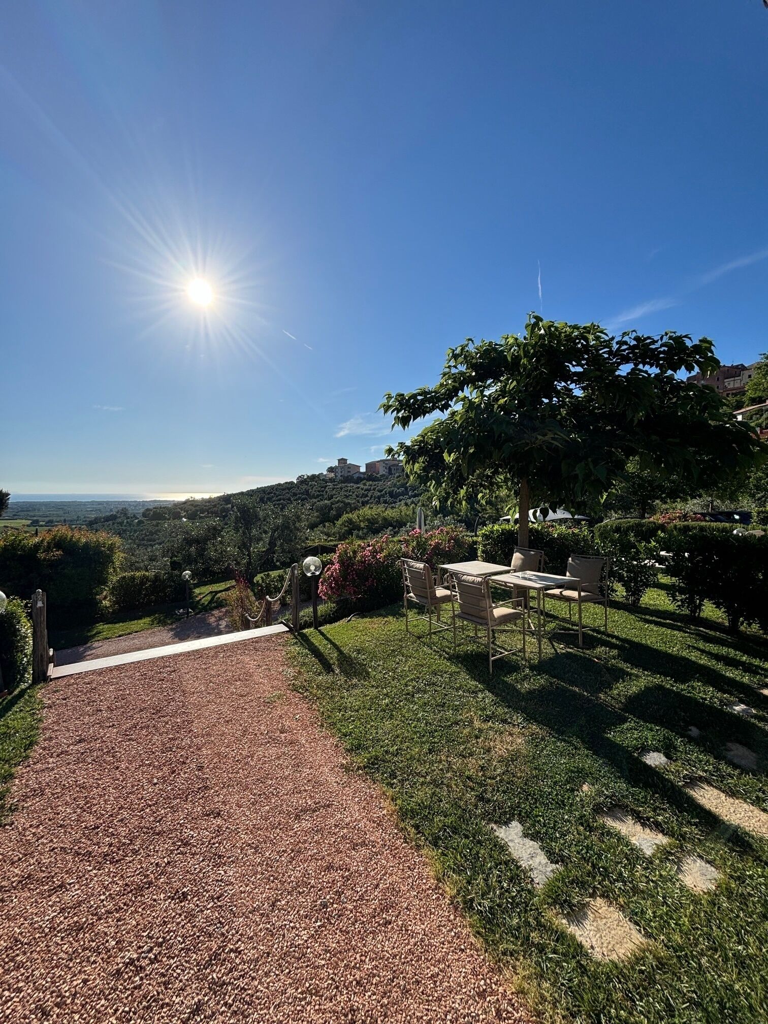Un patio jardín con un sendero de grava que conduce a una zona de estar, rodeado de abundante vegetación y vistas a edificios lejanos.