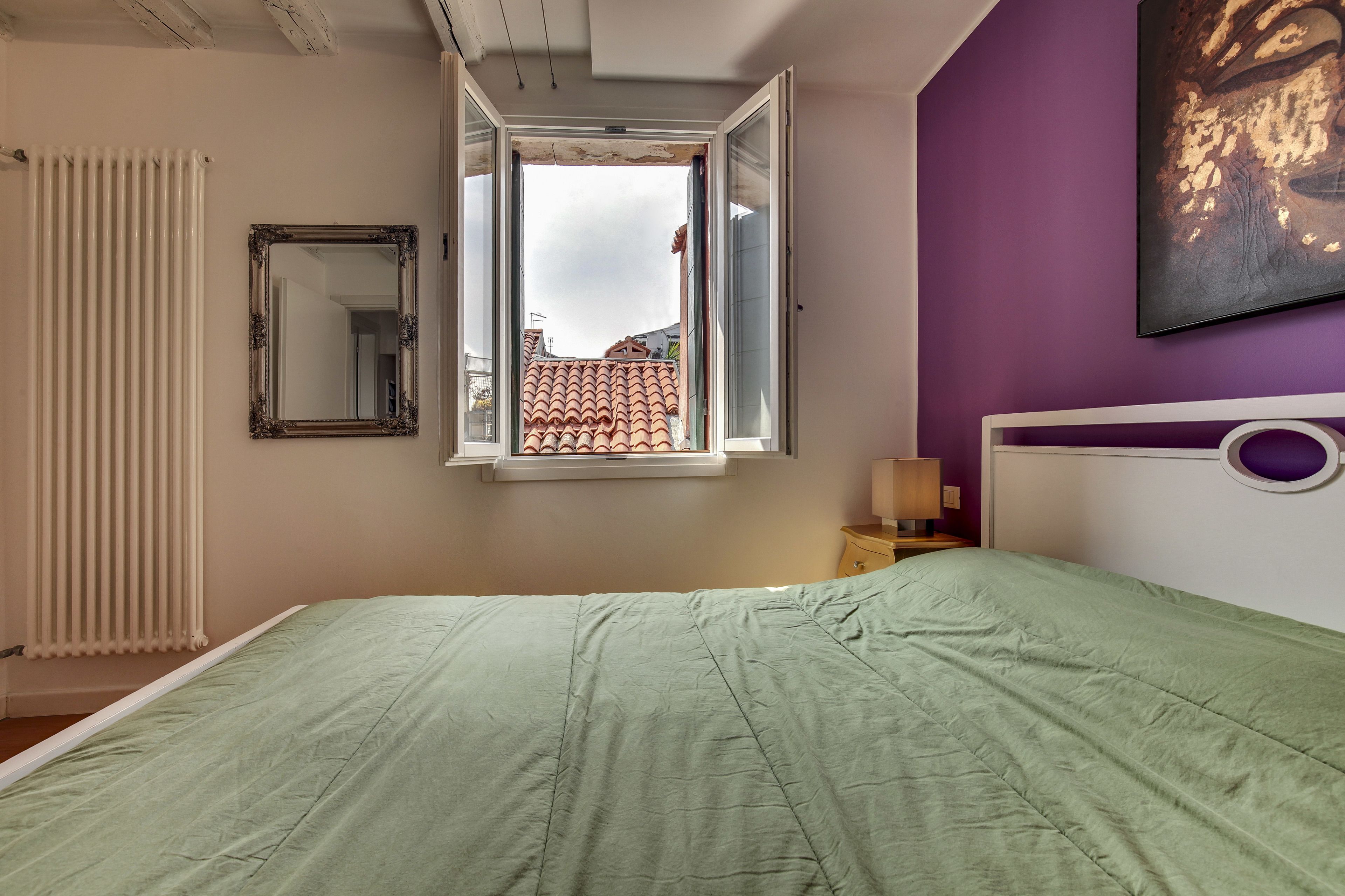 Une chambre avec une couvre-lit verte, un radiateur blanc, un miroir, une fenêtre donnant sur des immeubles et un mur violet.