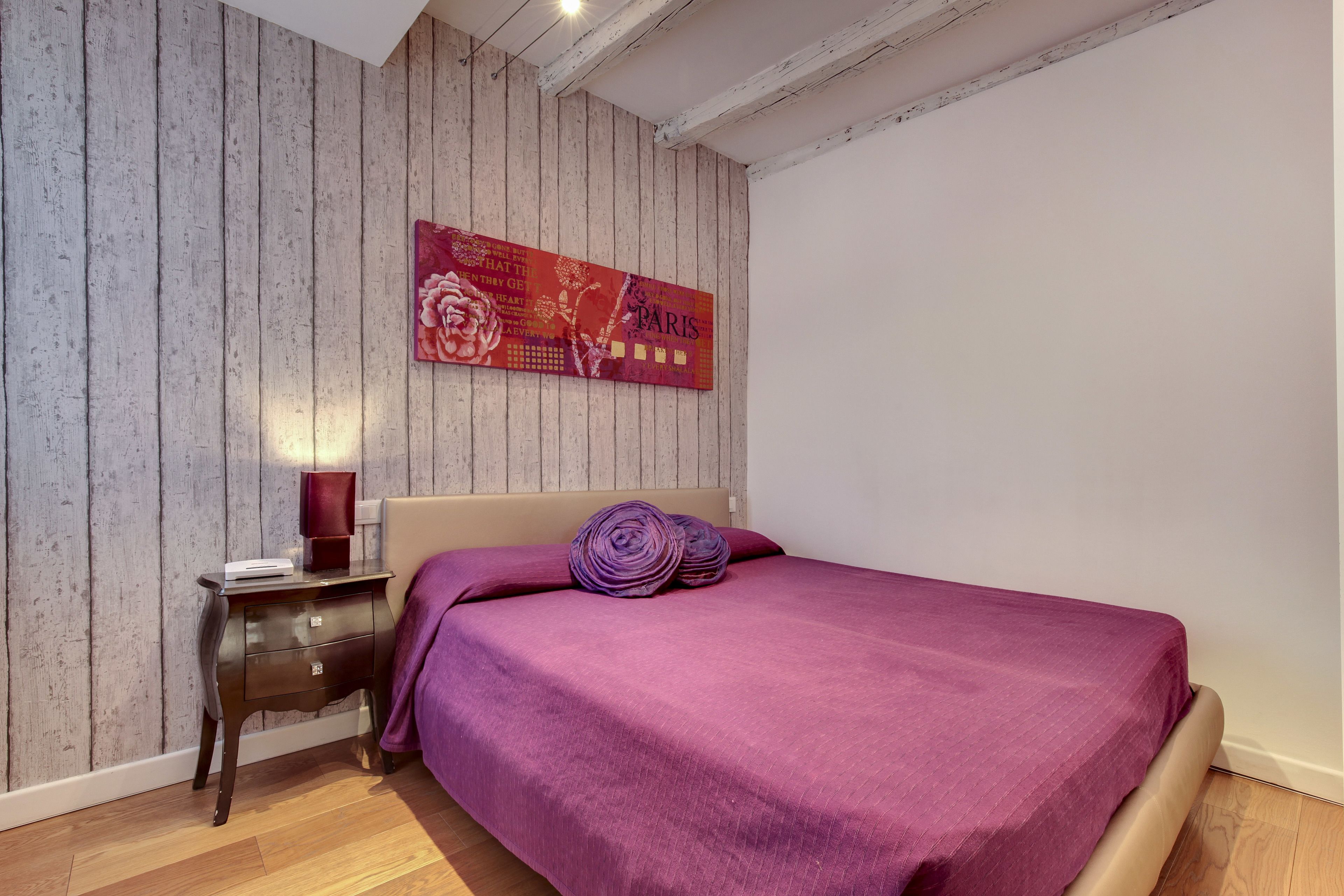 Une chambre avec un mur en bois, un lit recouvert d’une couverture violette et une table de chevet avec une lampe.