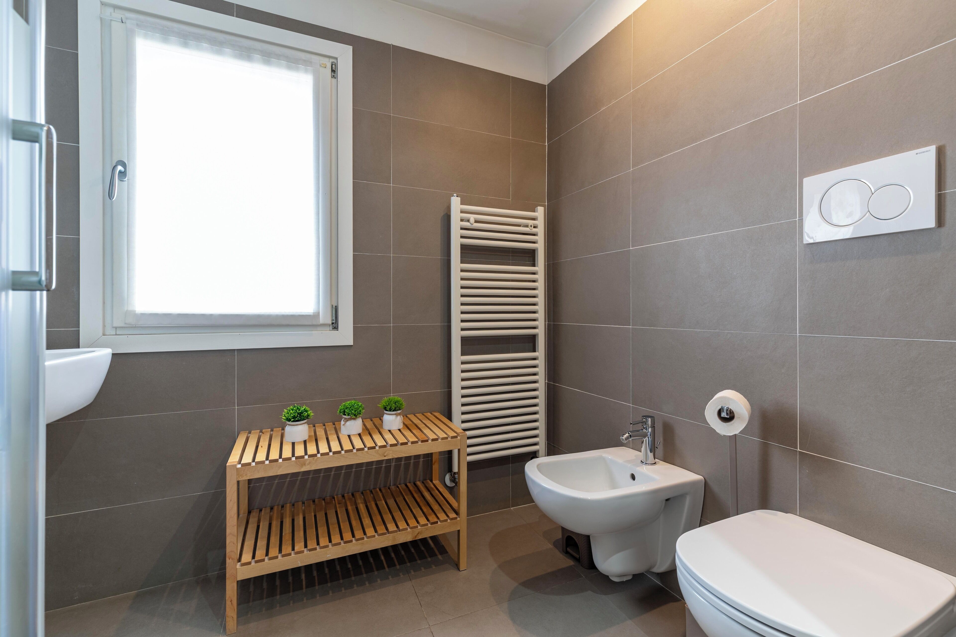 Un baño moderno con inodoro, bidé, estante de madera con plantas, un perchero para toallas y una ventana.