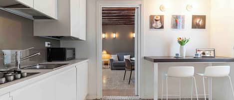 A modern kitchen with white cabinets, a black microwave, and a stainless steel sink. There is a bar area with two white stools and a vase of flowers. The room has a neutral color palette and a rustic ceiling.