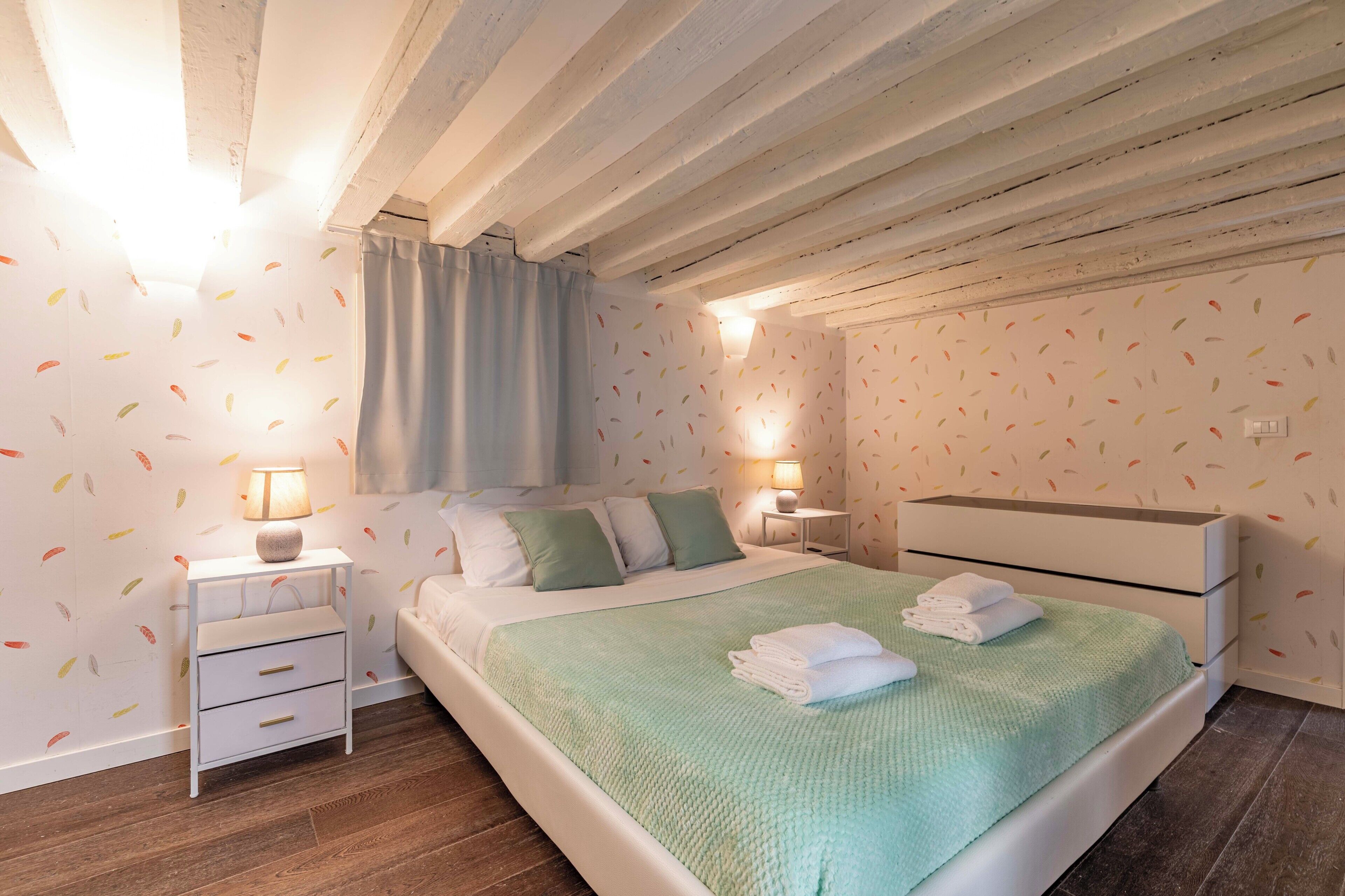 Une chambre à coucher comprenant un lit, des tables de chevet, une commode, une lampe, une fenêtre avec des rideaux et un plafond avec des poutres apparentes.