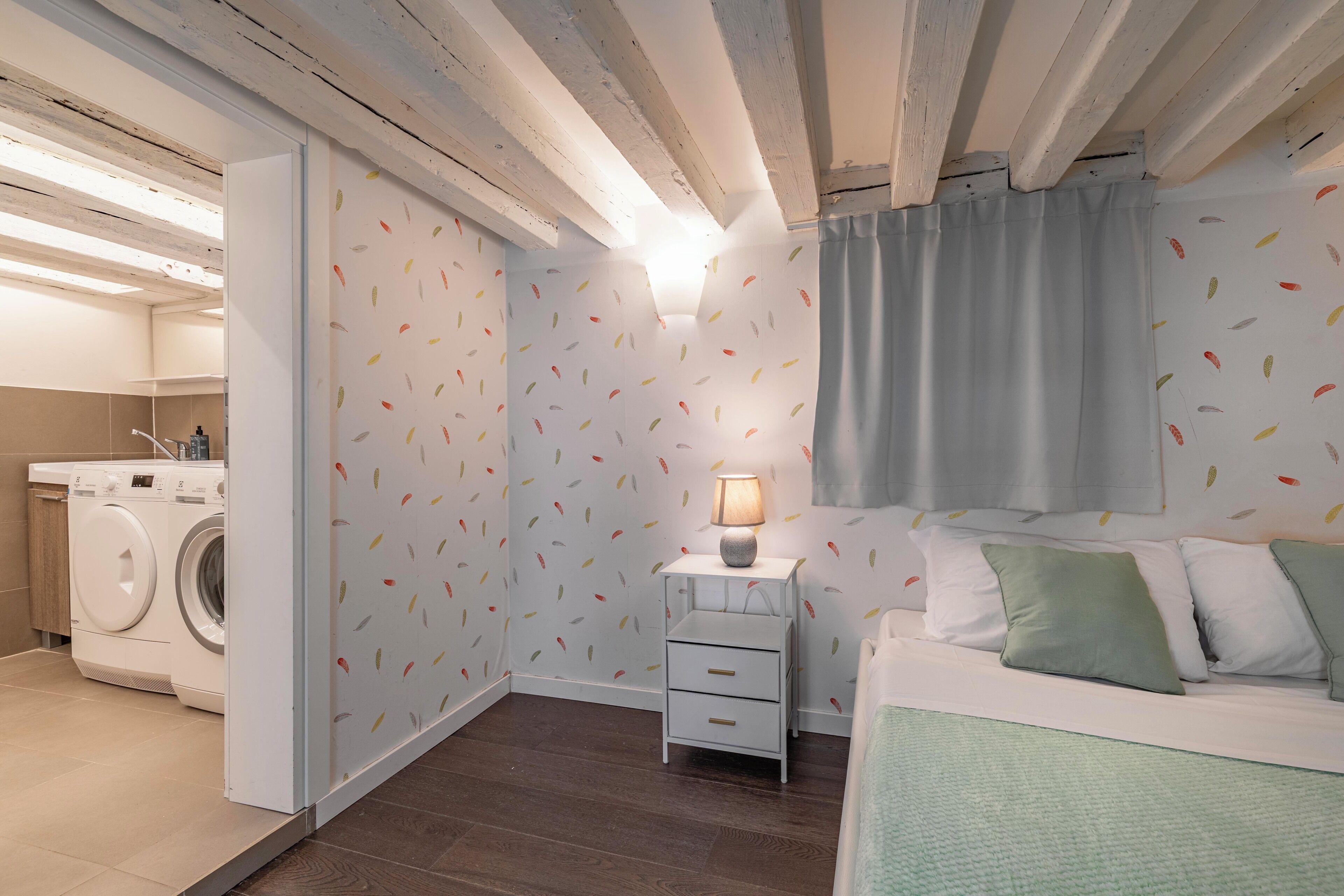 A compact bedroom with a washing machine, a bedside table with a lamp, and a bed with pillows.
