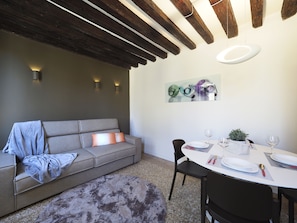 A modern living room with a grey sofa, a dining table set for two, and a ceiling with exposed wooden beams.
