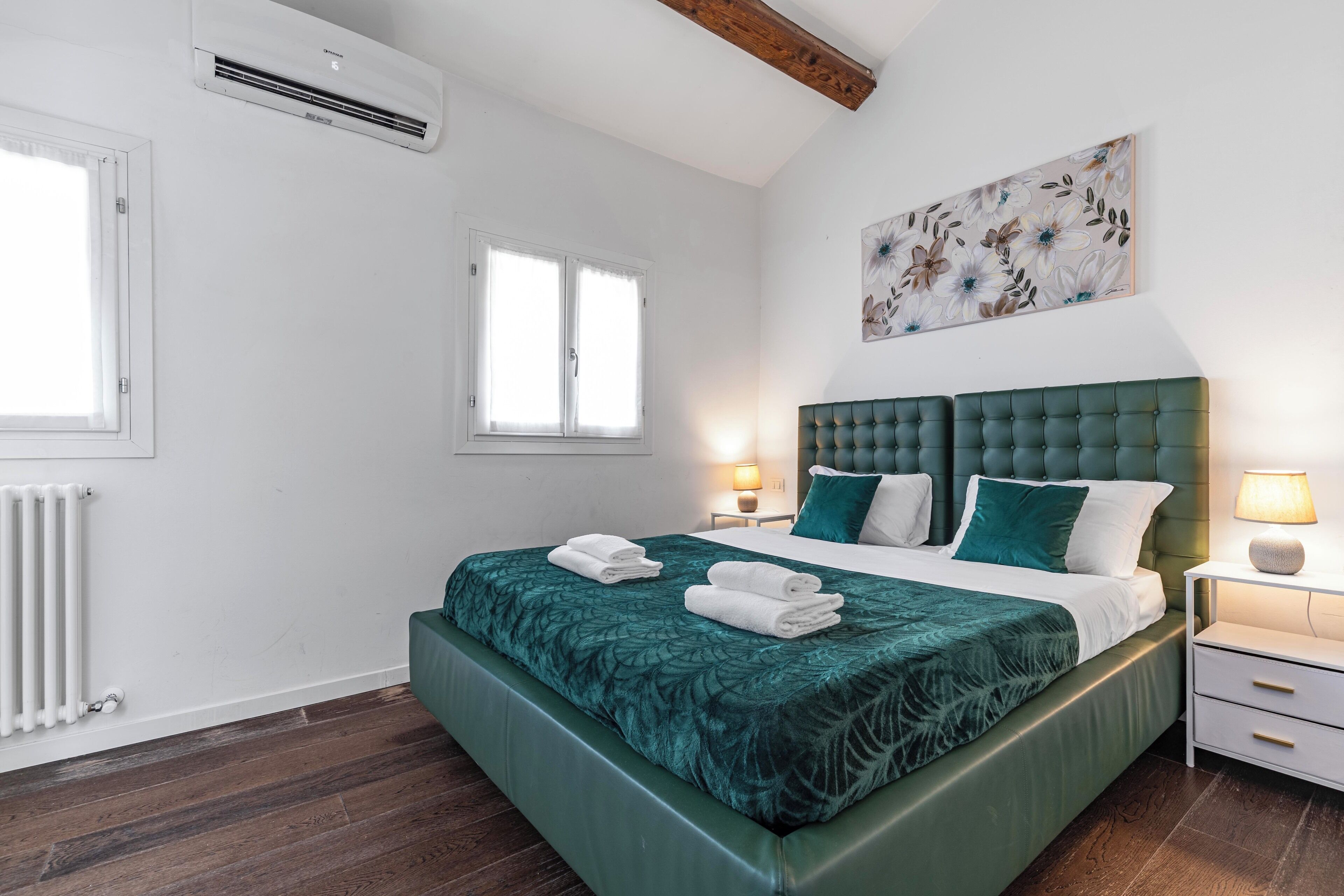 A bedroom with a green upholstered bed, white bedding, a radiator, a window, and a wall-mounted air conditioner.