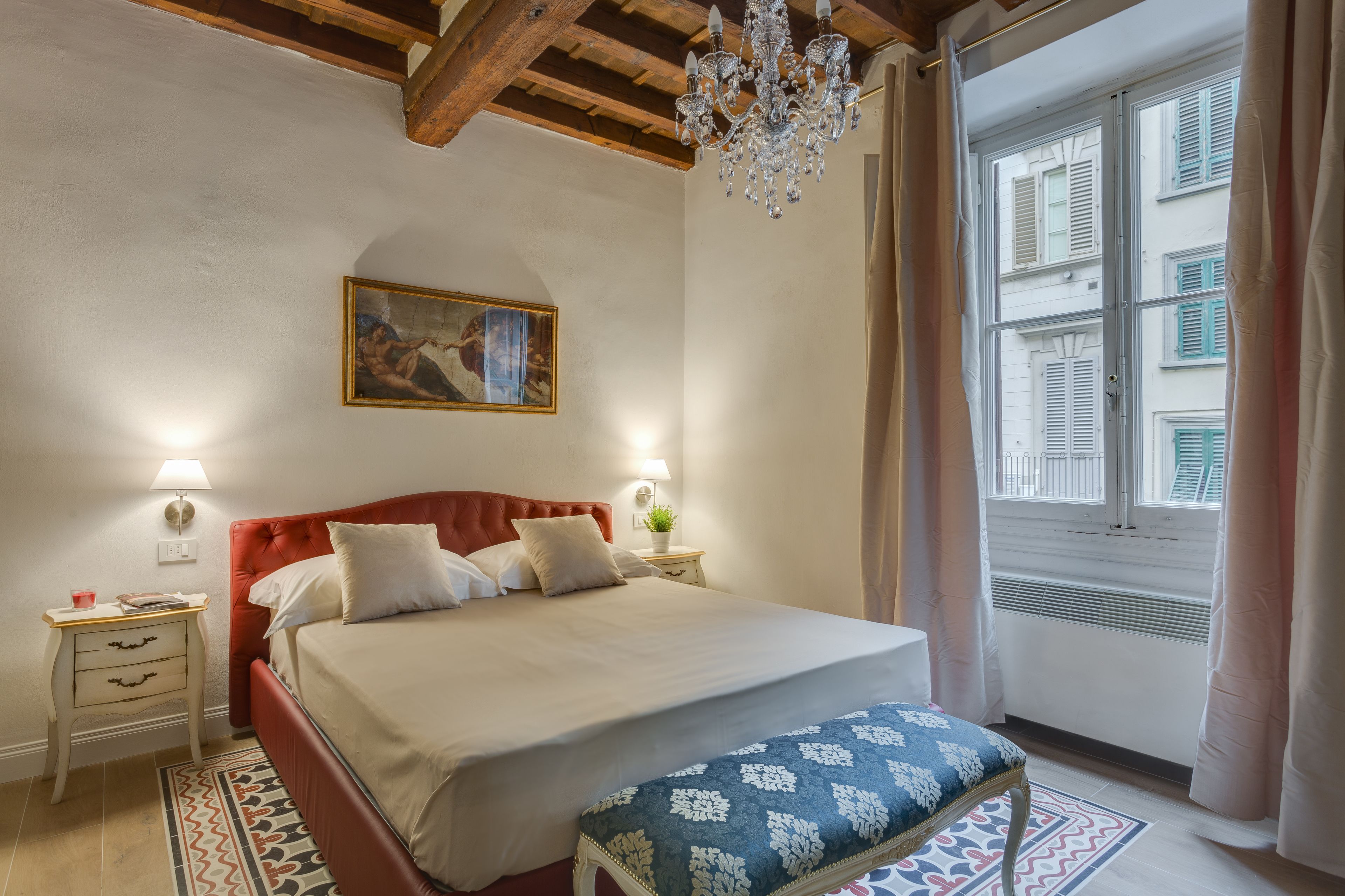 Une chambre à coucher avec un lit, des tables de chevet, un lustre, une fenêtre avec des rideaux et un tableau au mur.