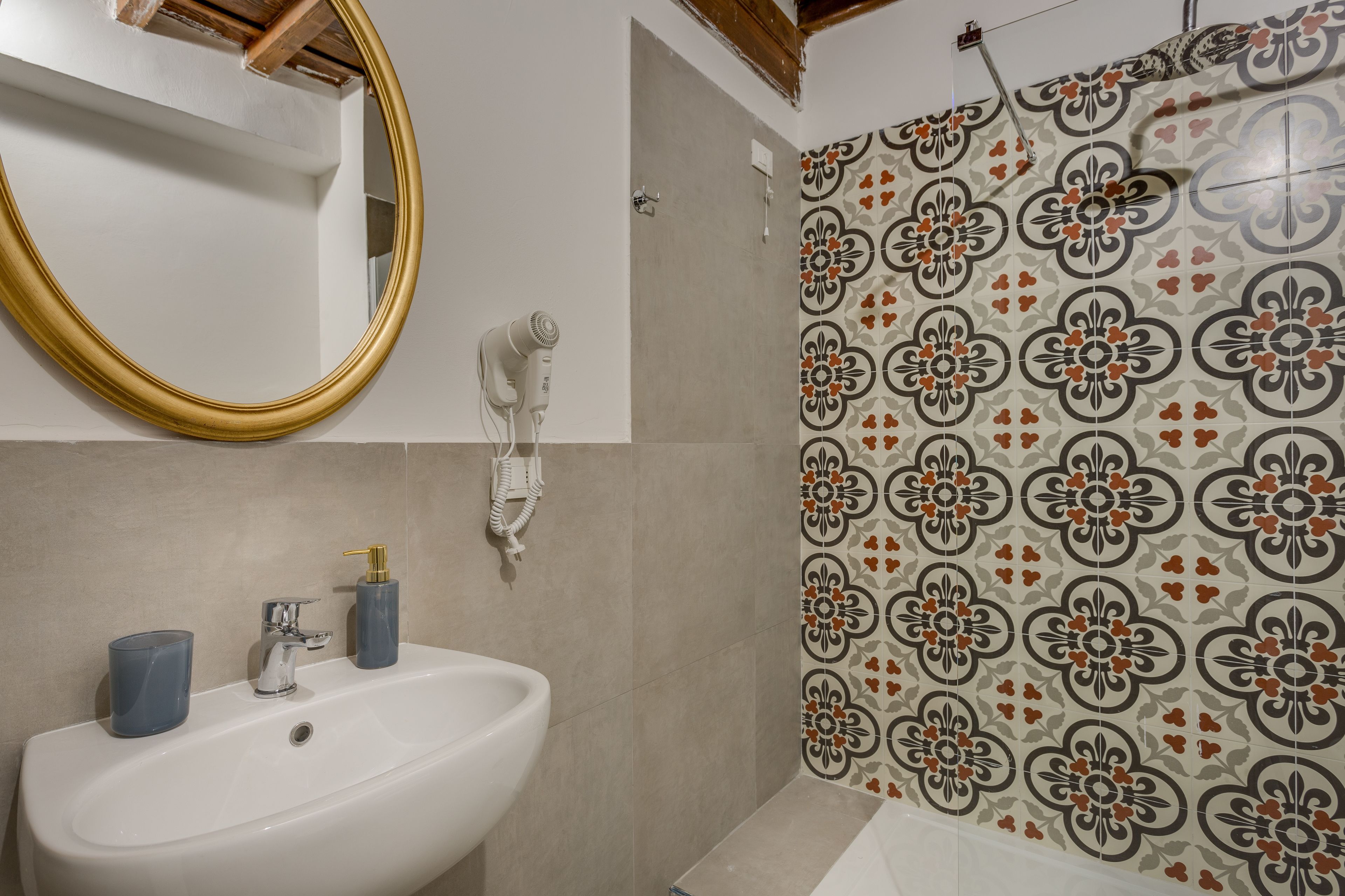 A bathroom with a white sink, a gold-framed mirror, a hairdryer, and a shower with patterned tiles.