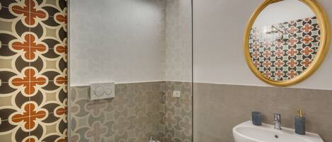 A bathroom with a patterned tile wall, a glass shower enclosure, a sink with a gold faucet, and a mirror with a gold frame.