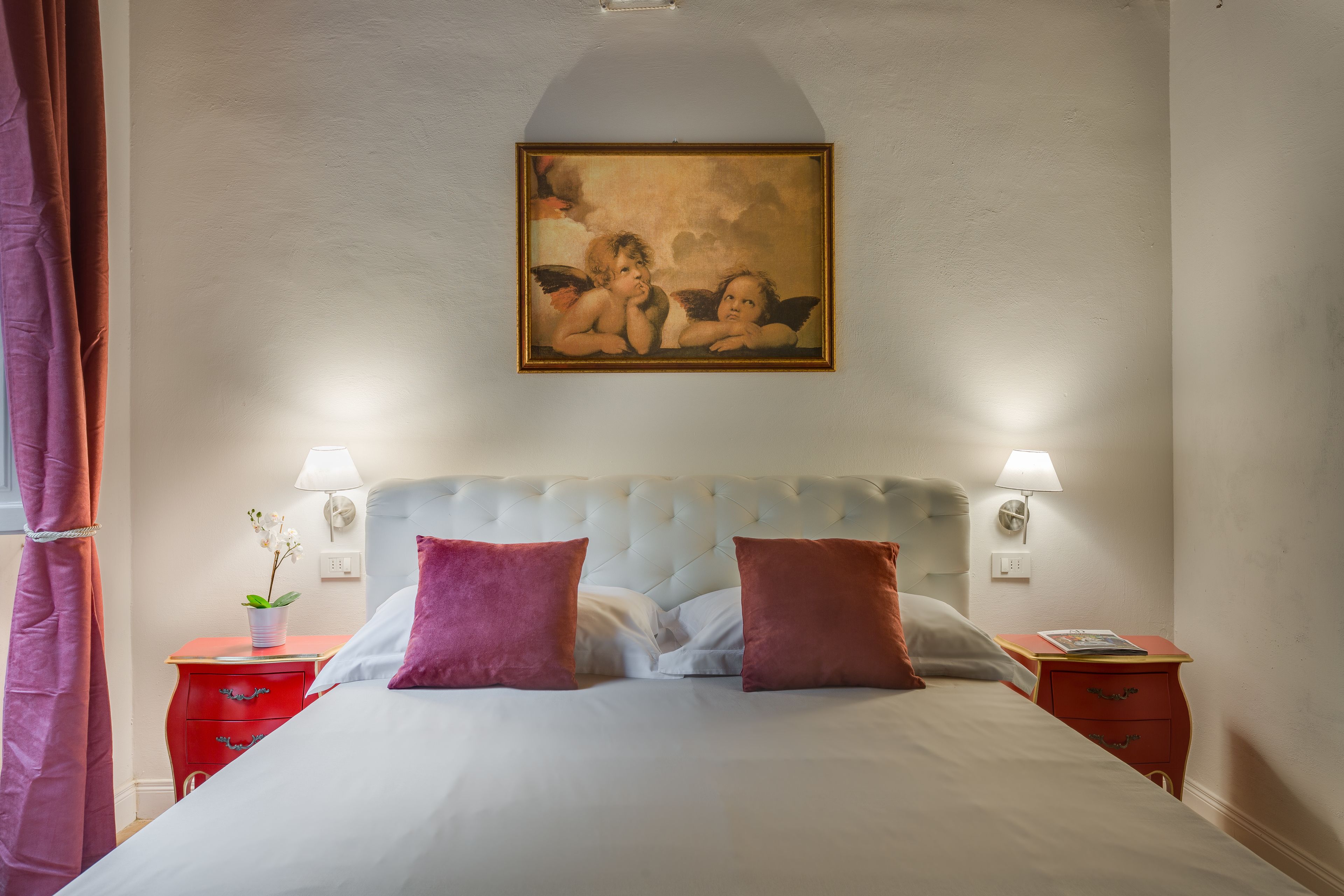 A neatly made bed with two pillows, a red nightstand with a lamp, and a painting above the bed.