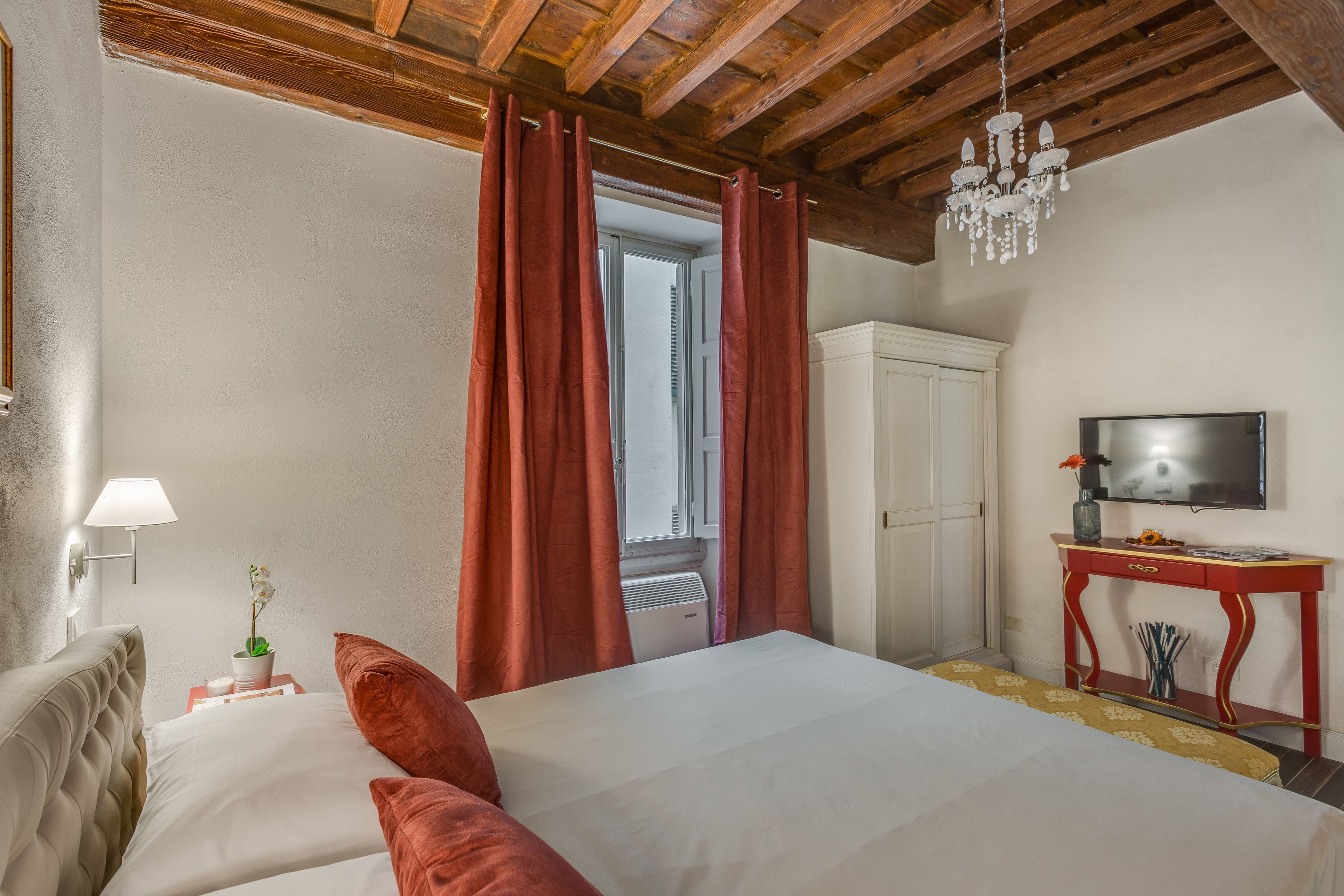A hotel room with a bed, a TV on a wooden console, a white wardrobe, and a window with red curtains.