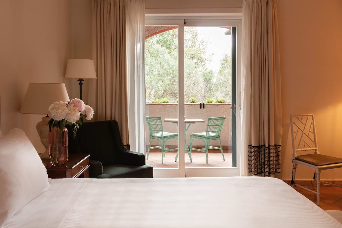 A hotel room with a bed, a desk with a lamp, a chair, a table with chairs, and a view of greenery through a sliding door.