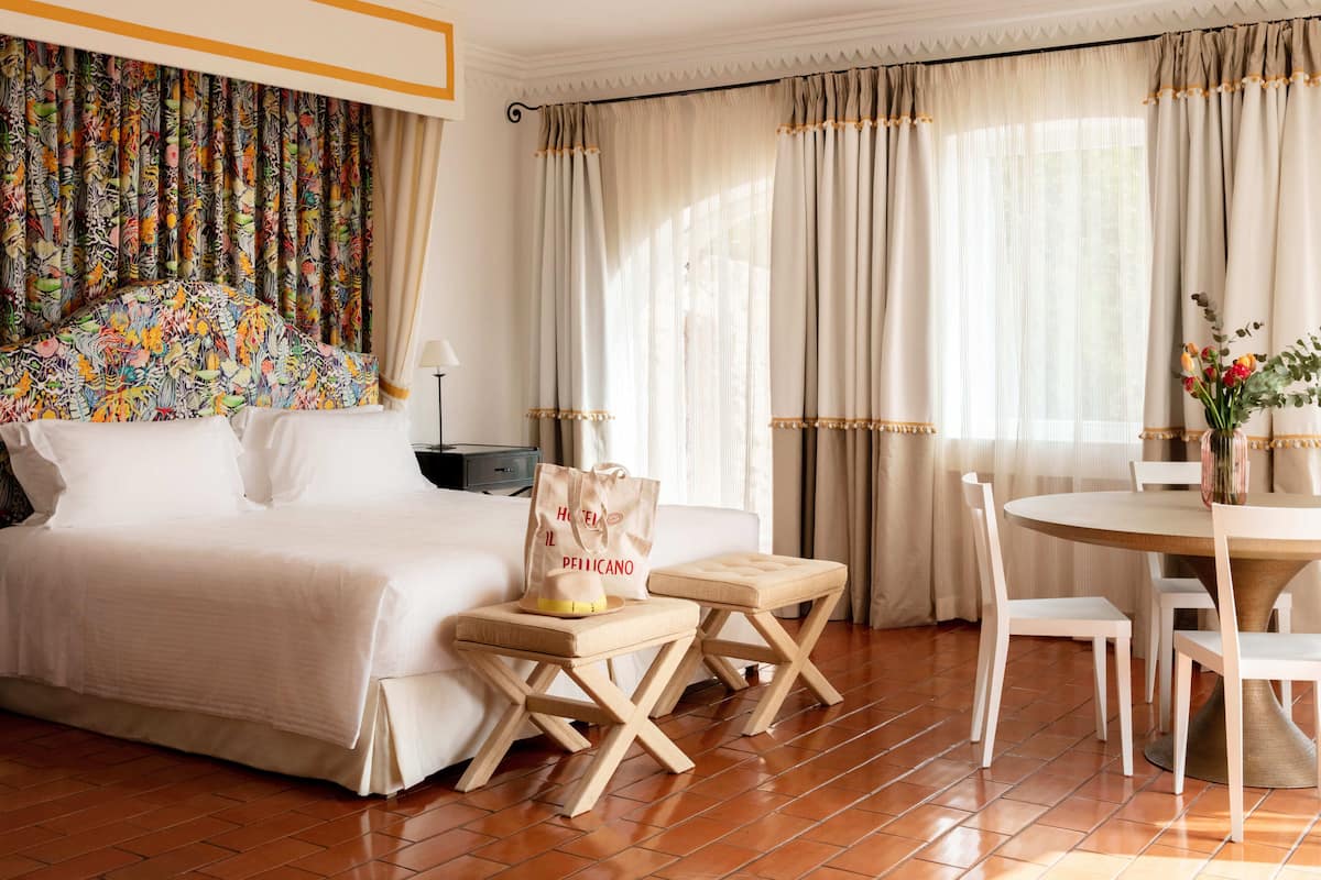 A hotel room with a bed, a table with chairs, a small stool, a bag, and curtains.