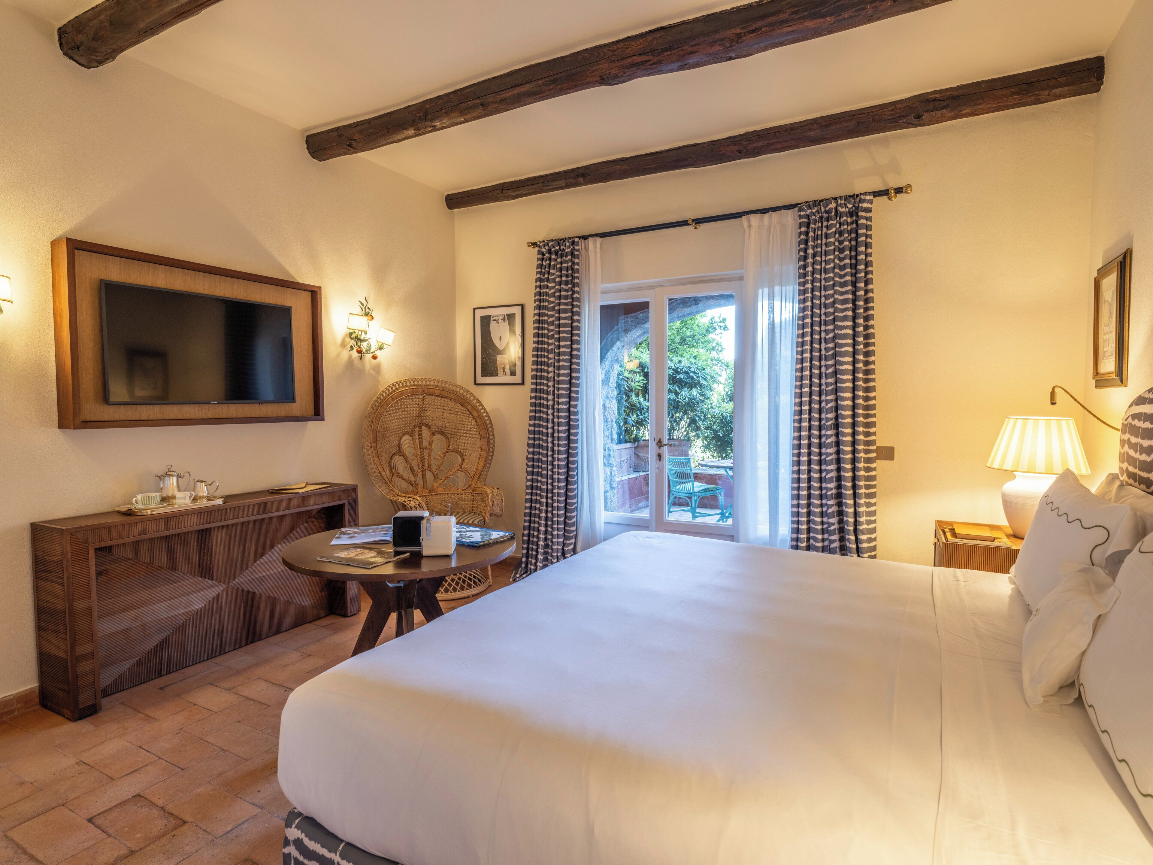 A bedroom with a large bed, a wooden headboard, a television, a wicker chair, a small table with a lamp, and a view through a glass door.