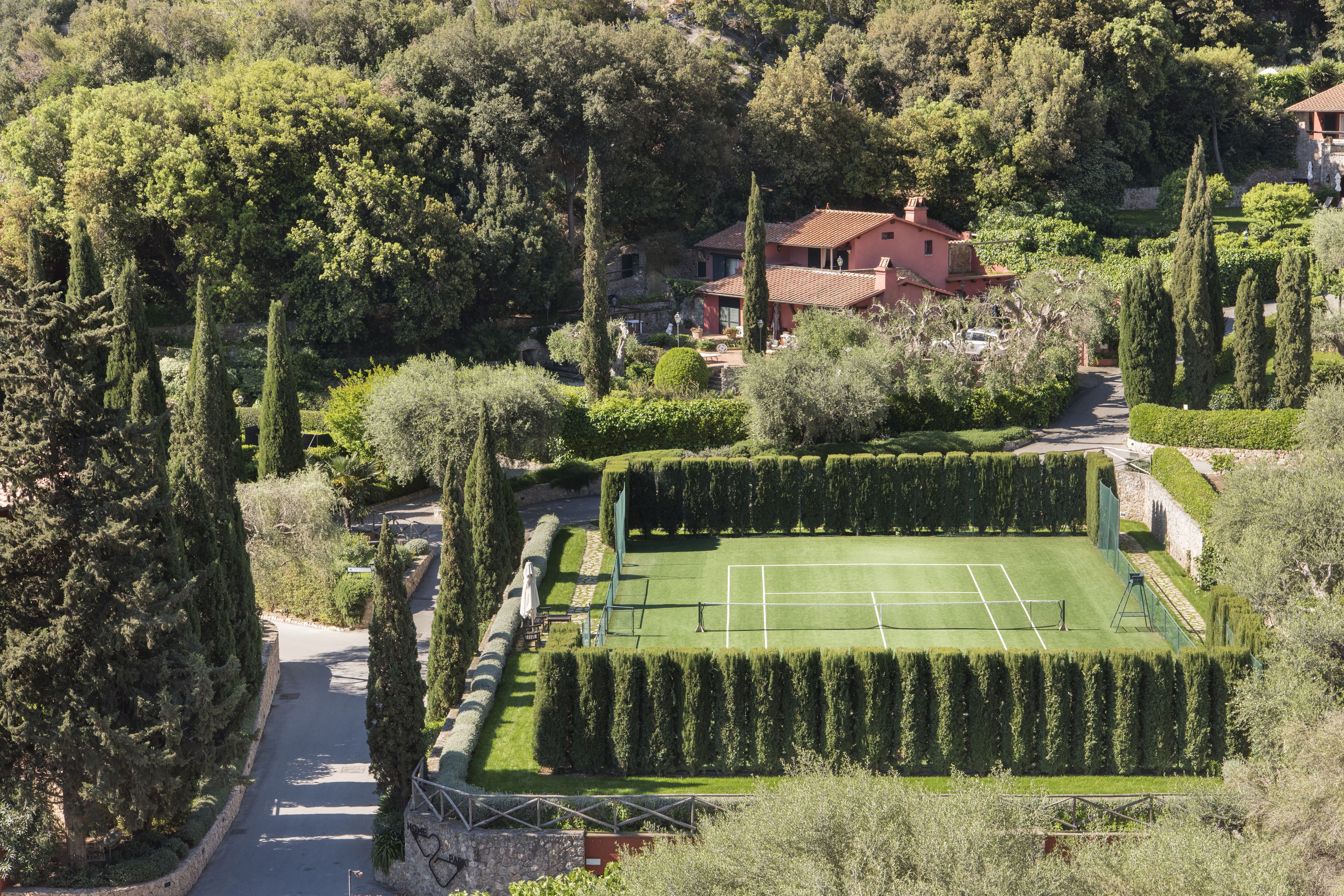 Una cancha de tenis rodeada de setos bien cuidados y altos árboles, con una casa al fondo.