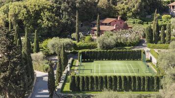 Una cancha de tenis rodeada de setos bien cuidados y altos árboles, con una casa al fondo.