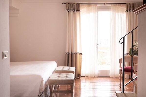 A bedroom with a bed, two ottomans, a staircase, and a view of the outdoors through sheer curtains.