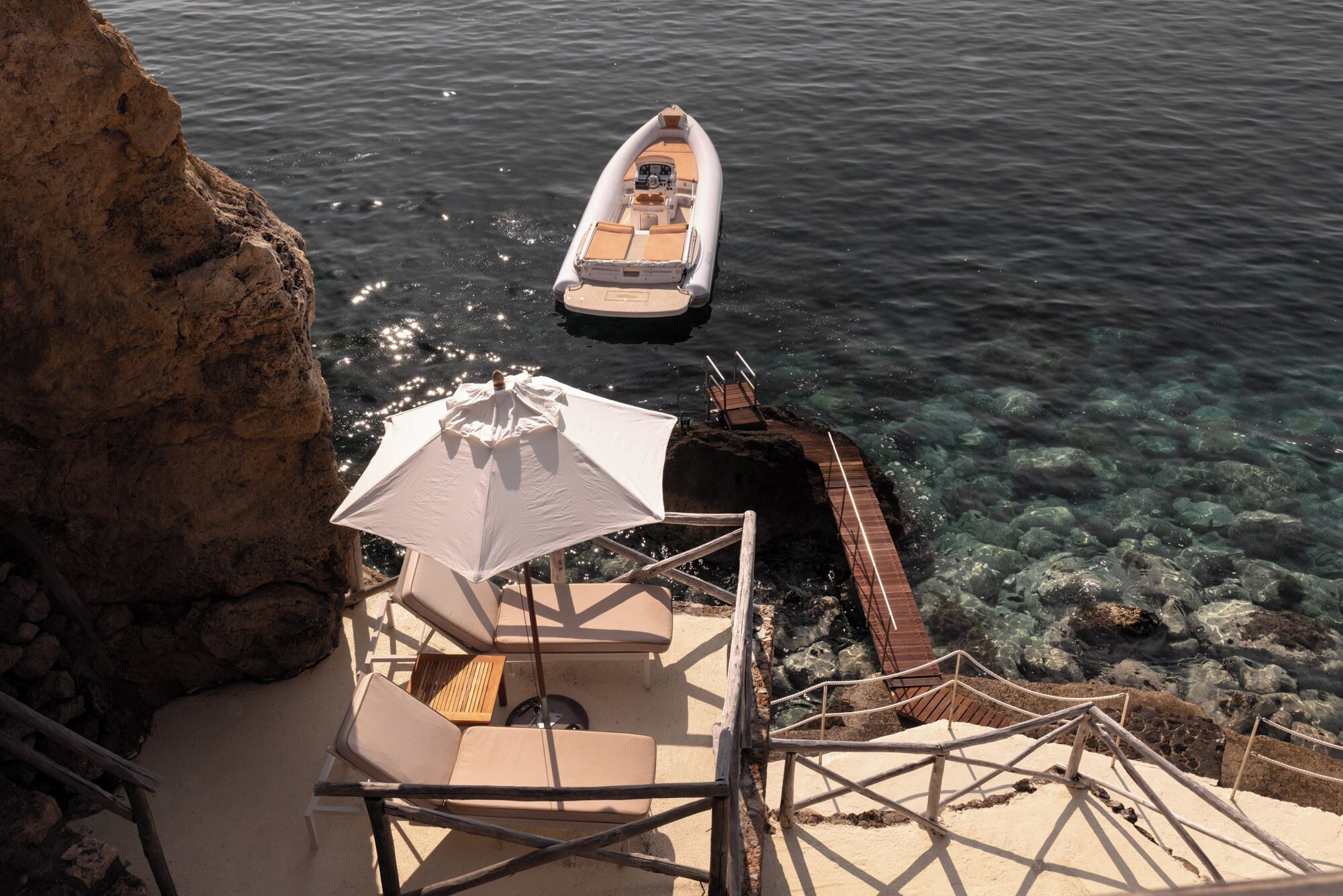 A boat docked near a rocky shore with clear water, a wooden deck, and a white umbrella.
