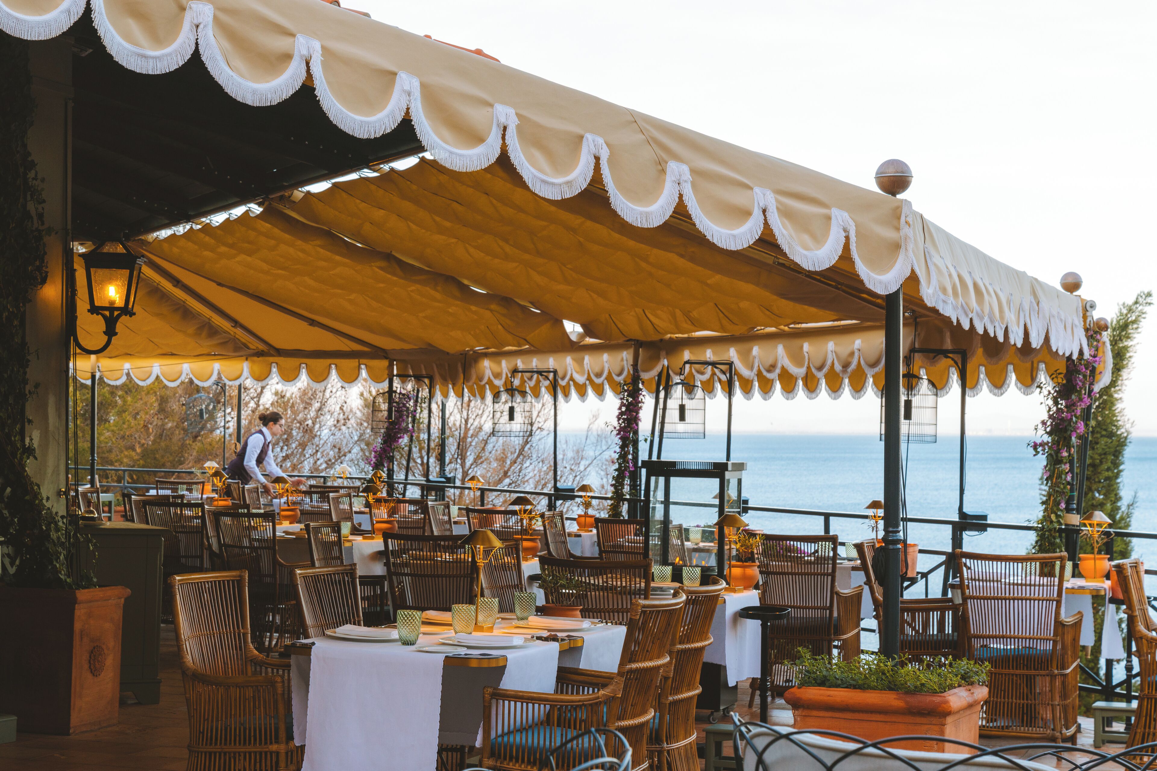 An outdoor dining area with tables and chairs, a covered section, and a view of the sea.