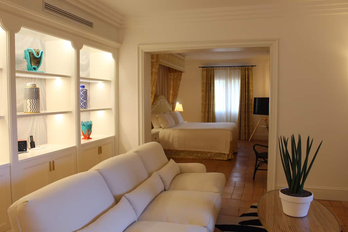 A hotel room with a white sofa, a bed with a gold headboard, a bedside table with a lamp, and a potted plant.
