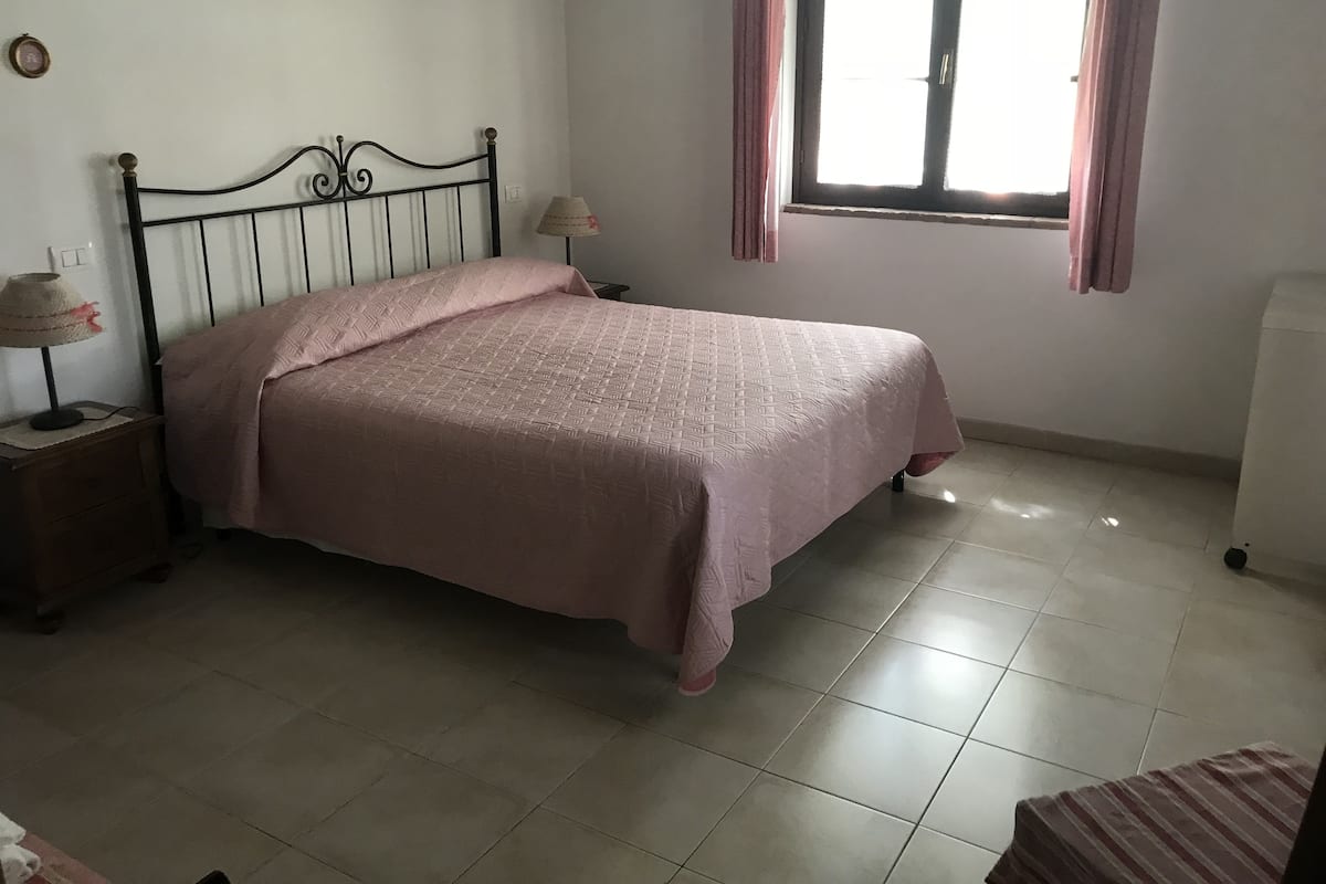 A bedroom with a bed, a nightstand, a lamp, a window with curtains, and a radiator.