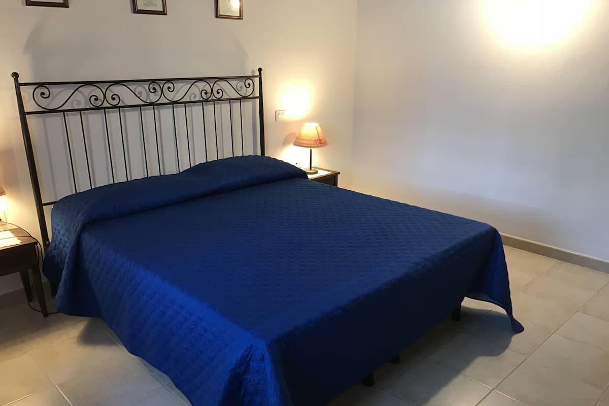 A bed with a blue bedspread, a metal headboard, a bedside table with a lamp, and a tiled floor.