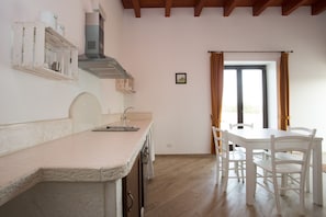 A kitchen with a white countertop, wooden cabinets, and a dining area with white chairs and a table. - Masseria Stella (Gioia del Colle)