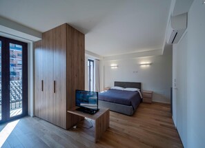 A modern bedroom with a wooden wardrobe, a bed with grey bedding, a TV on a wooden stand, and a balcony with a view of buildings.