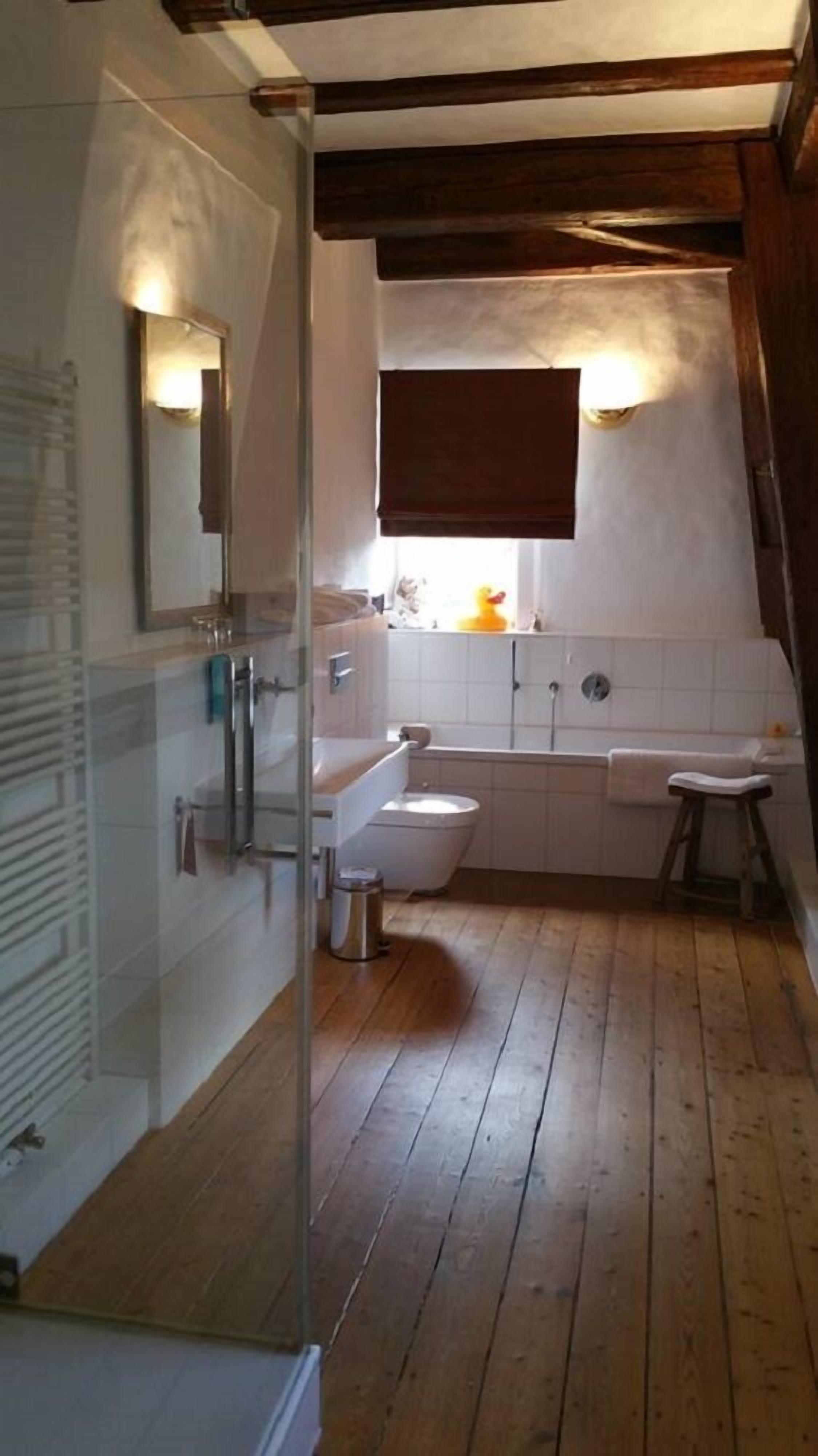 Un baño con piso de madera, azulejos blancos, un inodoro, un lavabo y un espejo.
