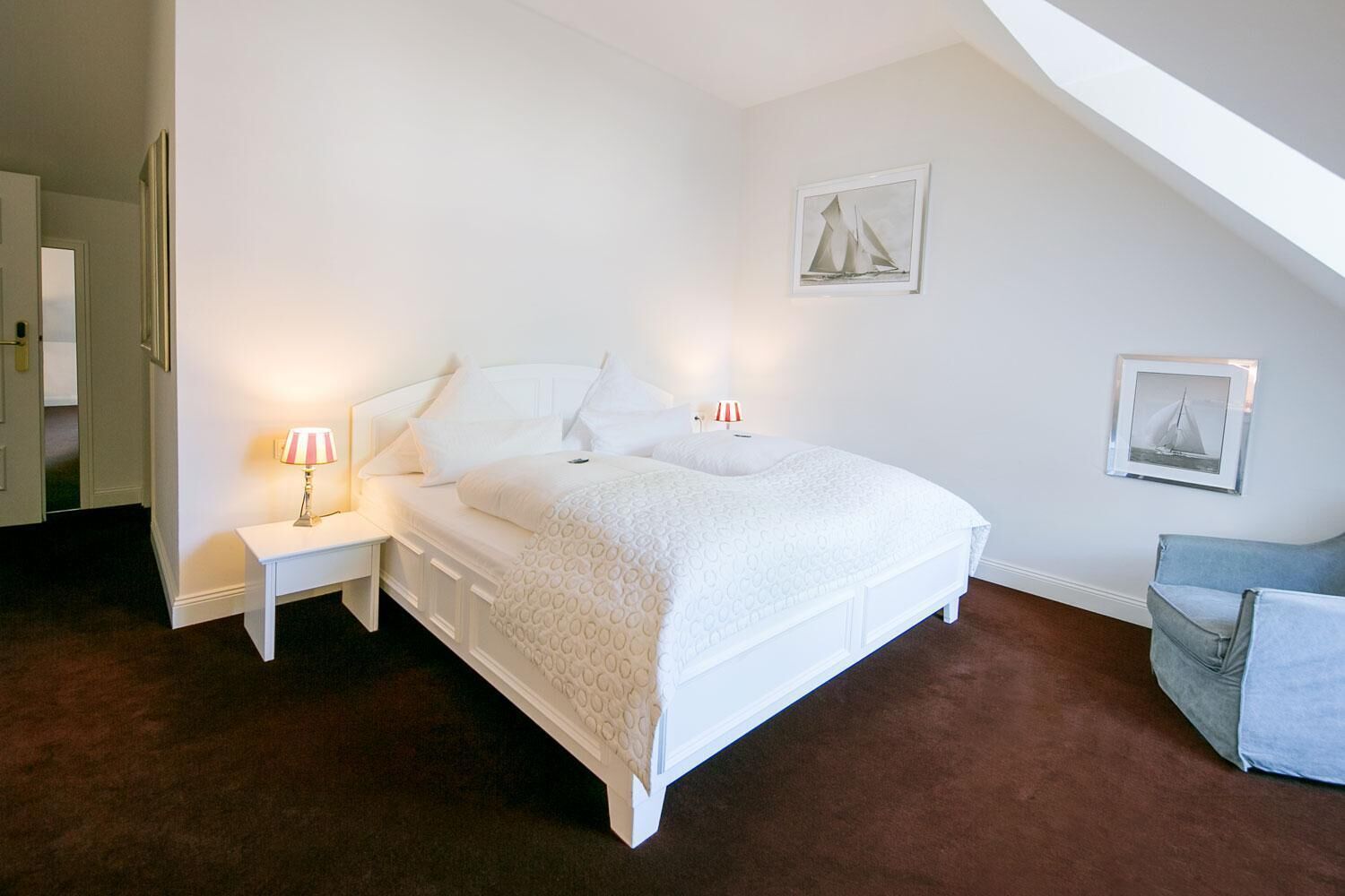Une chambre à coucher avec un lit blanc, deux tables de chevet avec des lampes, une chaise et deux tableaux encadrés accrochés au mur.